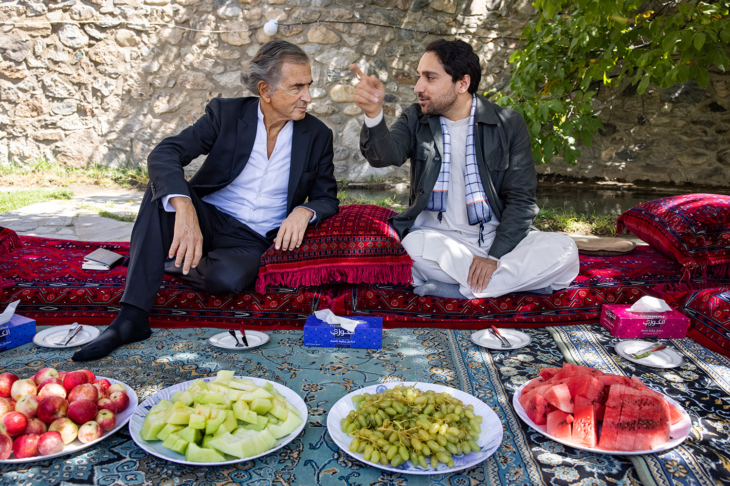 Bernard-Henri Lévy et Ahmad Massoud assis sur des tapis dans un jardin devant des saladiers de fruits
