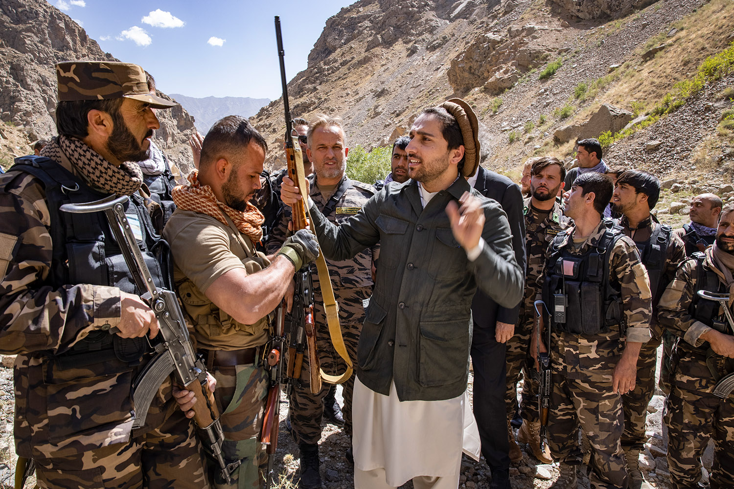 Ahmad Massoud porte un fusil, au milieu des montagnes de pierres, il est entouré par ses hommes en tenues militaires.