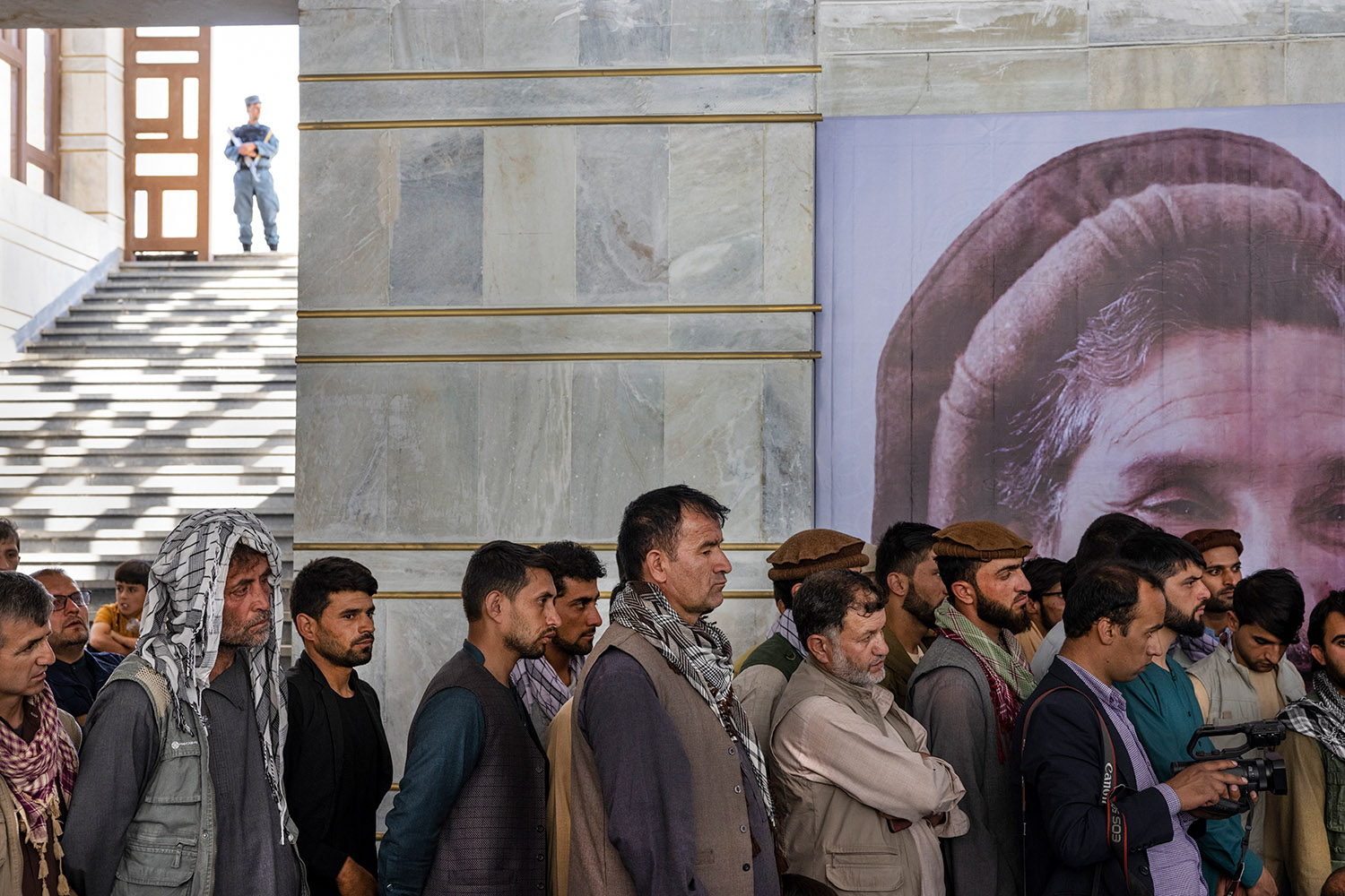 Une file d'hommes attendent, devant un grand portait du Commandant Massoud.