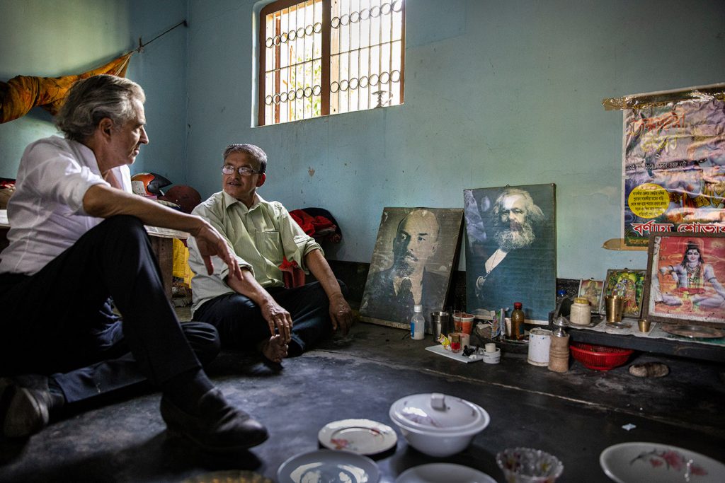 Bernard-Henri Lévy dans la chambre de sa jeunesse à Dacca, assis par terre, face à des portraits de Lénine et Marx et des icônes religieuses indiennes.