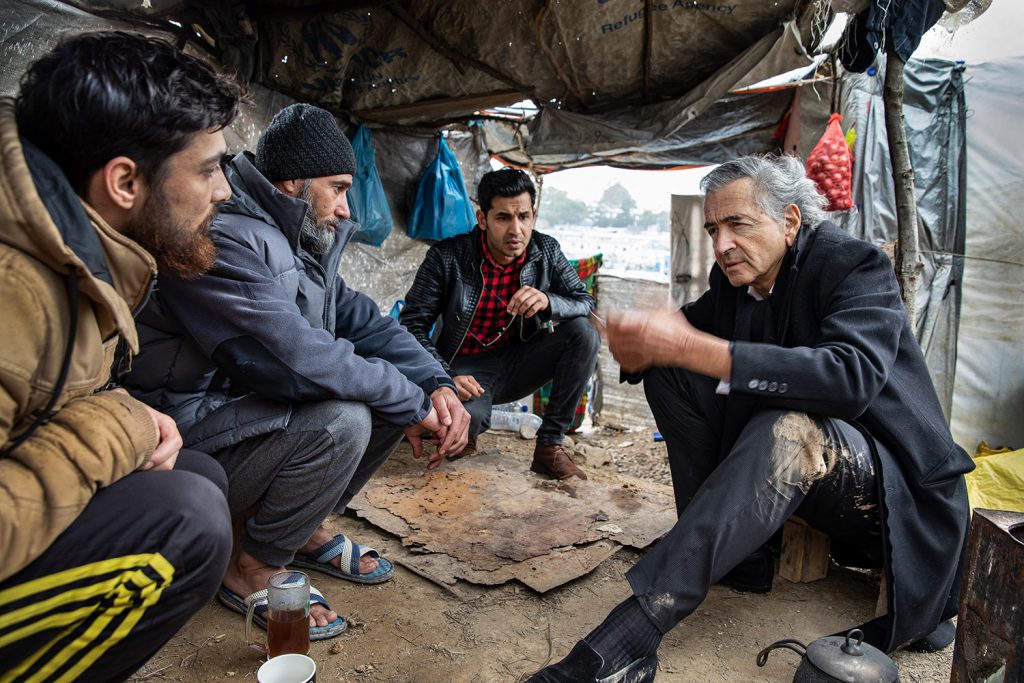 Bernard-Henri Lévy parle avec trois hommes, ils sont accroupis dans la boue sous une tente.