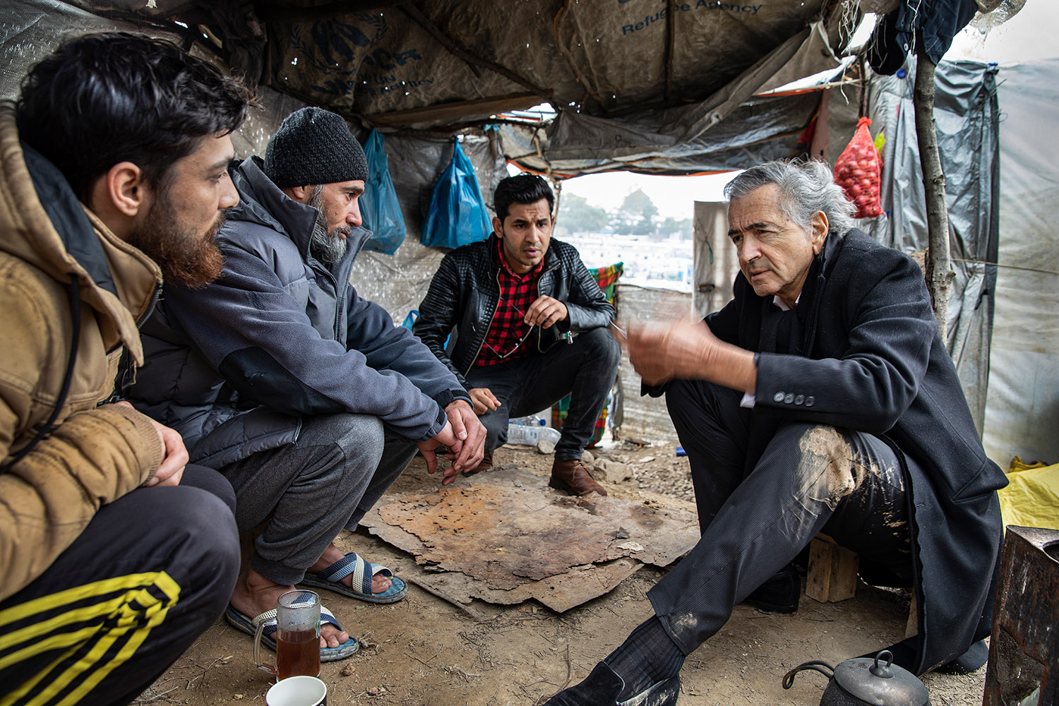 Bernard-Henri Lévy parle avec trois hommes, ils sont accroupis dans la boue sous une tente.
