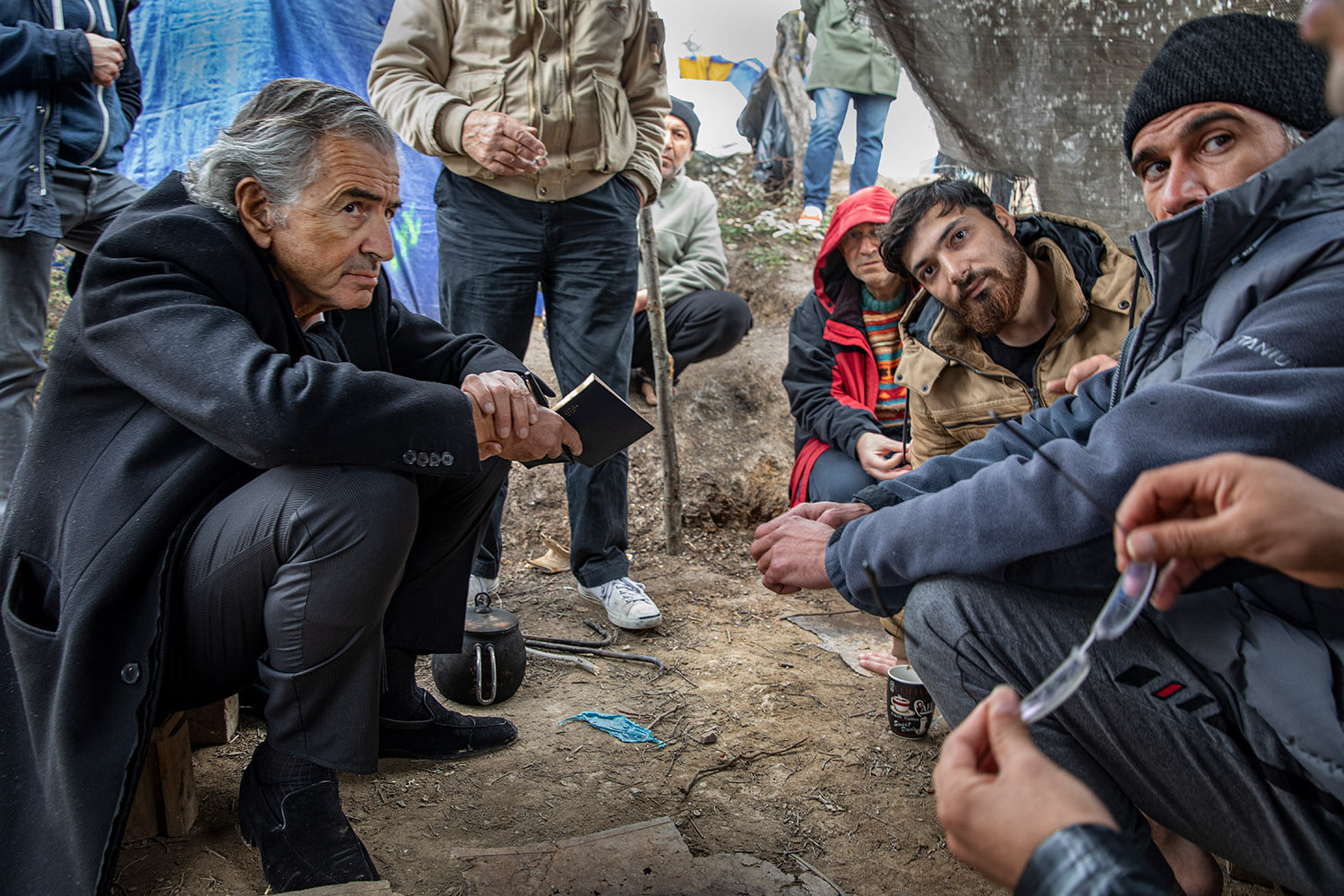 Bernard-Henri Lévy écoute des migrants. Il tient un stylo et un carnet. Tout le monde est accroupit dans la boue.