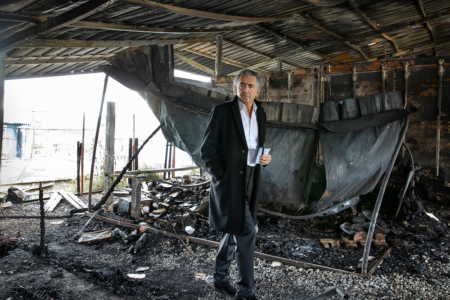 Bernard-Henri Lévy marche, un carnet à la main, dans les décombres d'un incendie dans le camp de migrants de Moria.