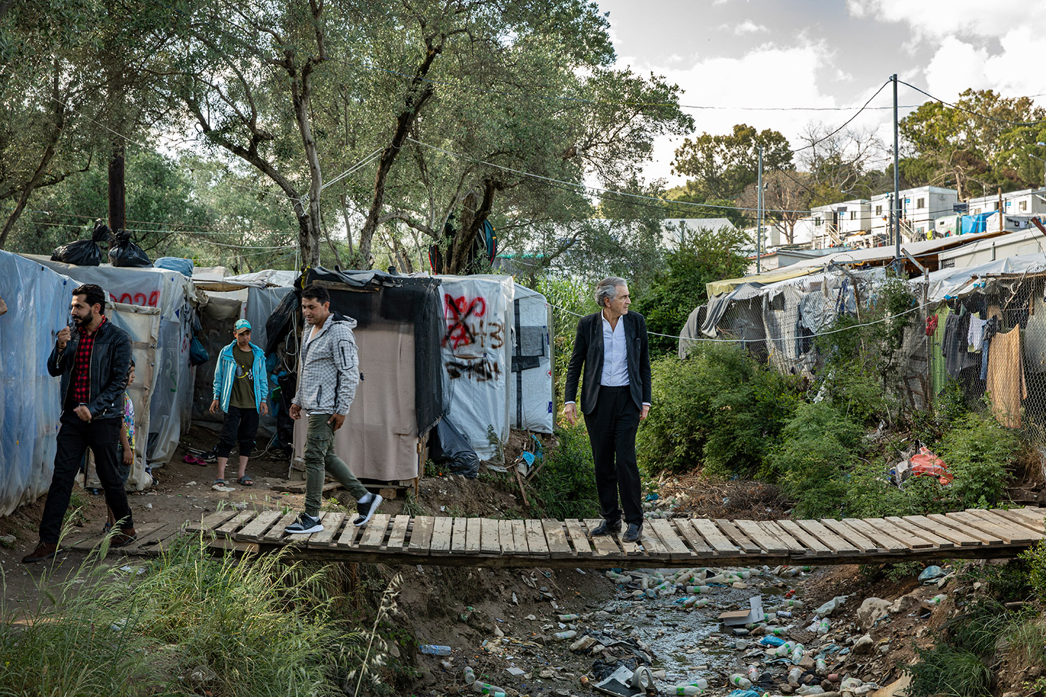 Bernard-Henri Lévy sur une passerelle de Moria qui passe au dessus des déchets et des écoulements d'eaux sales, à proximité des tentes des réfugiés.