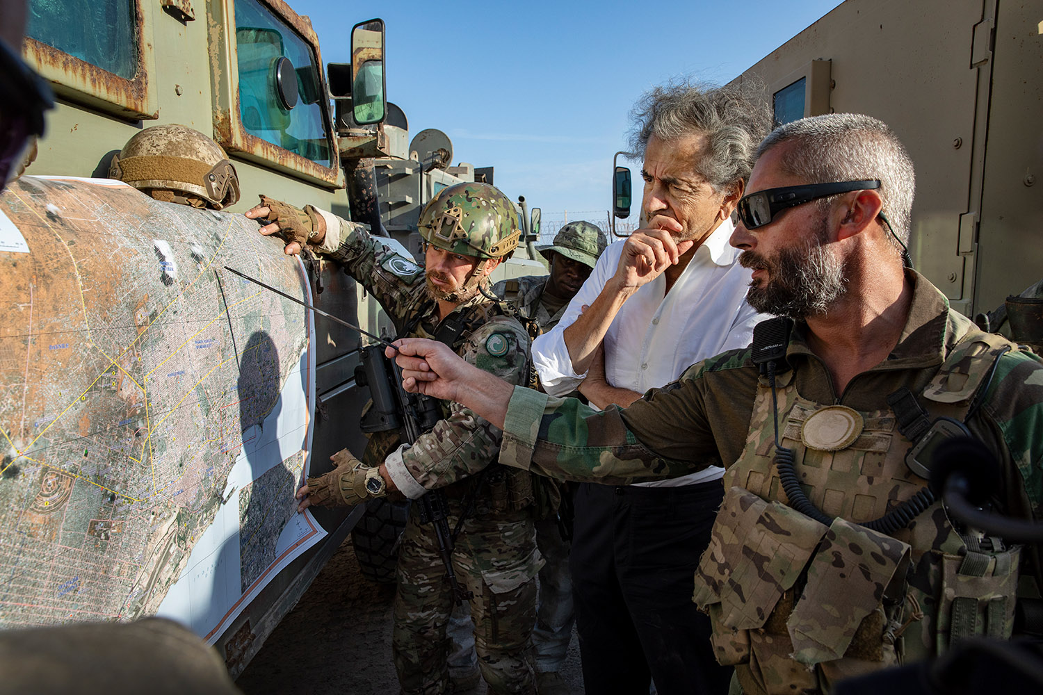 Bernard-Henri Lévy assiste à la préparation d’une intervention contre les milices islamistes, avec des militaires de Bancroft. L'un d'eux désigne un point sur une carte tendue sur la portière d'un véhicule blindé.