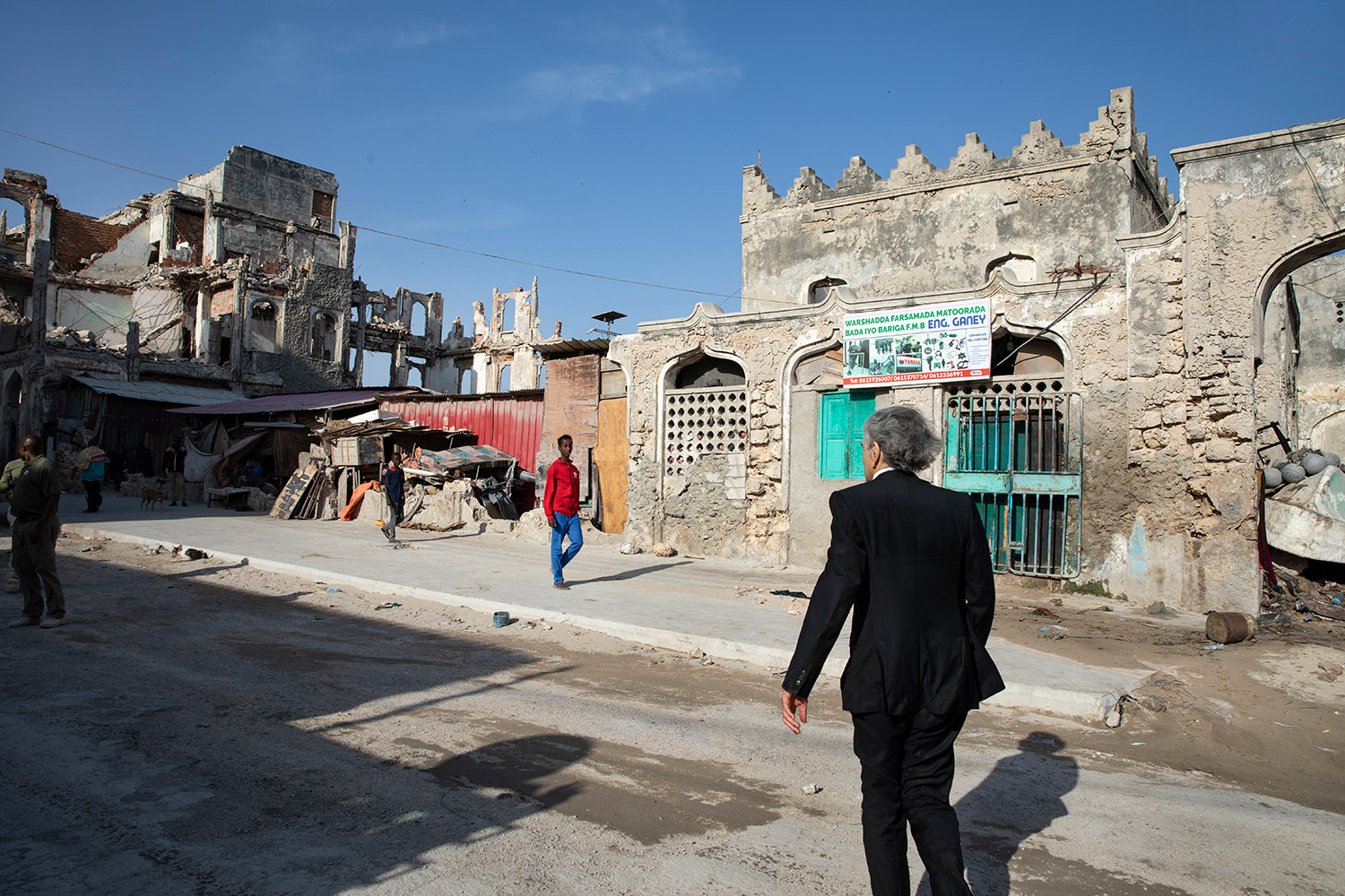 Bernard-Henri Lévy à Mogadiscio, il marche de dos au milieu de bâtiments à l'abandon ou en ruines.