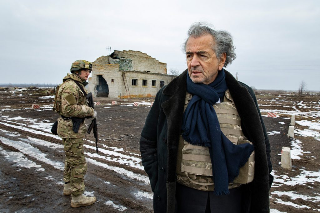 Bernard-Henri Lévy à Shyrokyne. A gauche on voit un militaire en uniforme, et au fond une maison détruite. Il y a de la neige au sol.