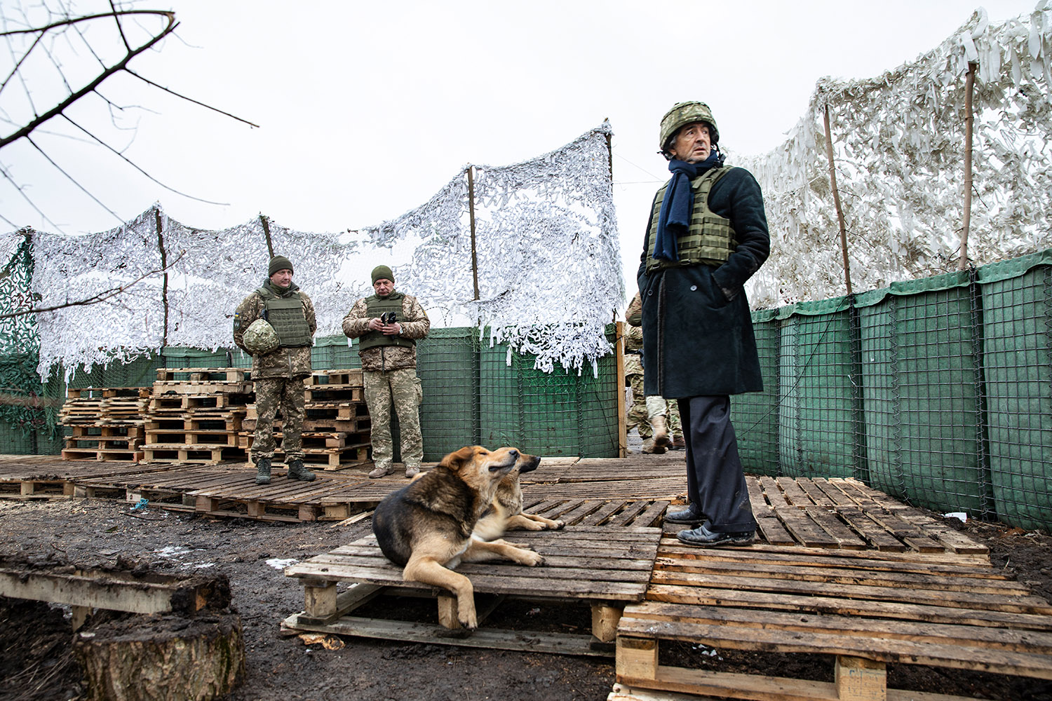 Bernard-Henri Lévy debout sur des palettes de bois, il porte un casque militaire et un gilet par balle sur son manteau. À ses pieds il y a un chien, et au fond à gauche, deux militaires.