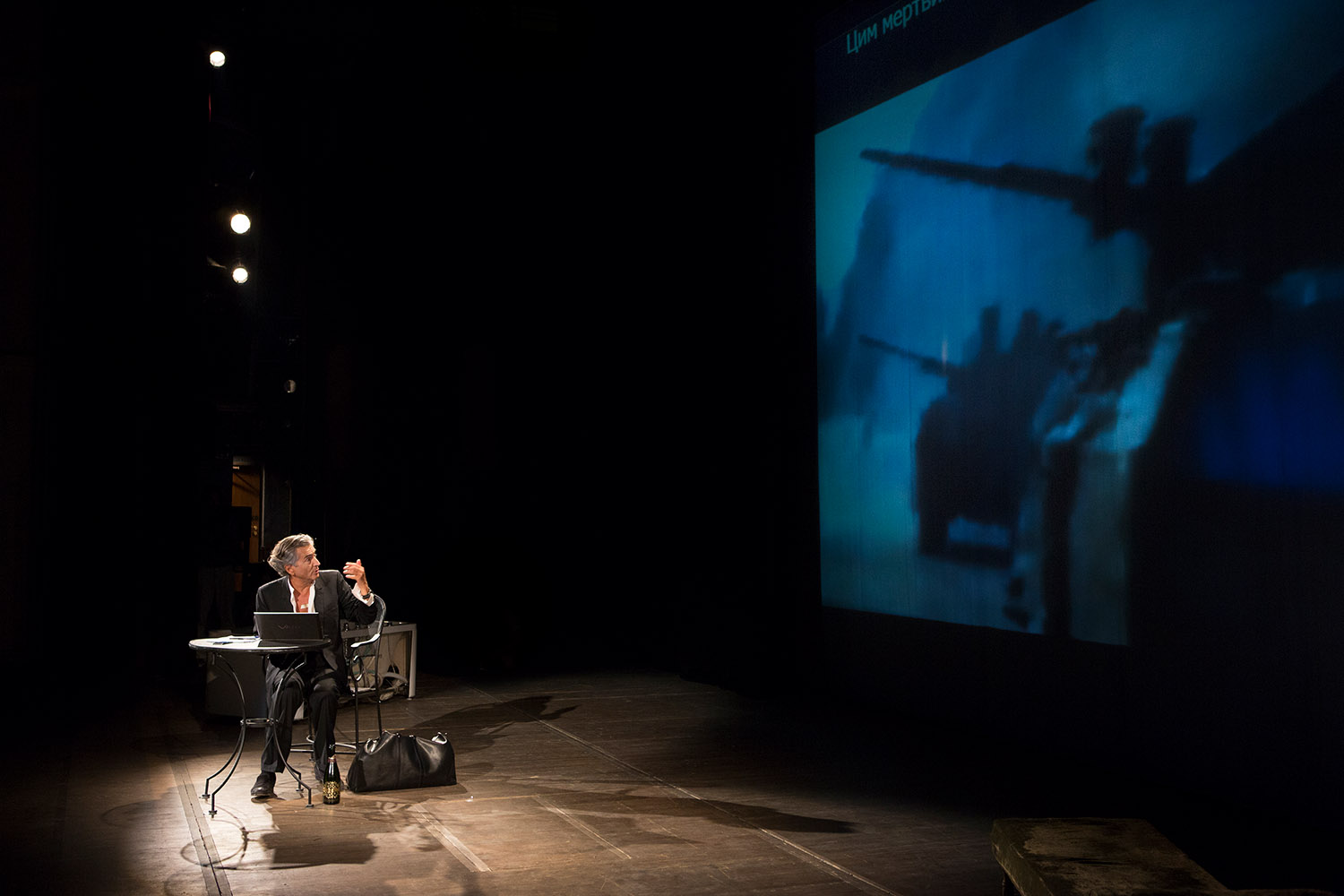 Bernard-Henri Lévy interprète sa pièce « Hôtel Europe » à l'Opéra d'Odessa. Il regarde, assis, le fond de la scène où est projeté des images de combats militaires avec des chars.