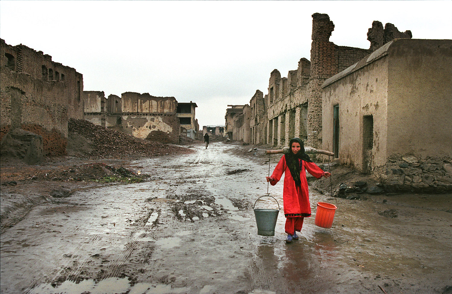 Dans une rue de Kaboul, le sol est boueux et les maisons sont détruites, probablement par les Talibans. Une femme afghane porte des seaux d'eau sur ses épaules.