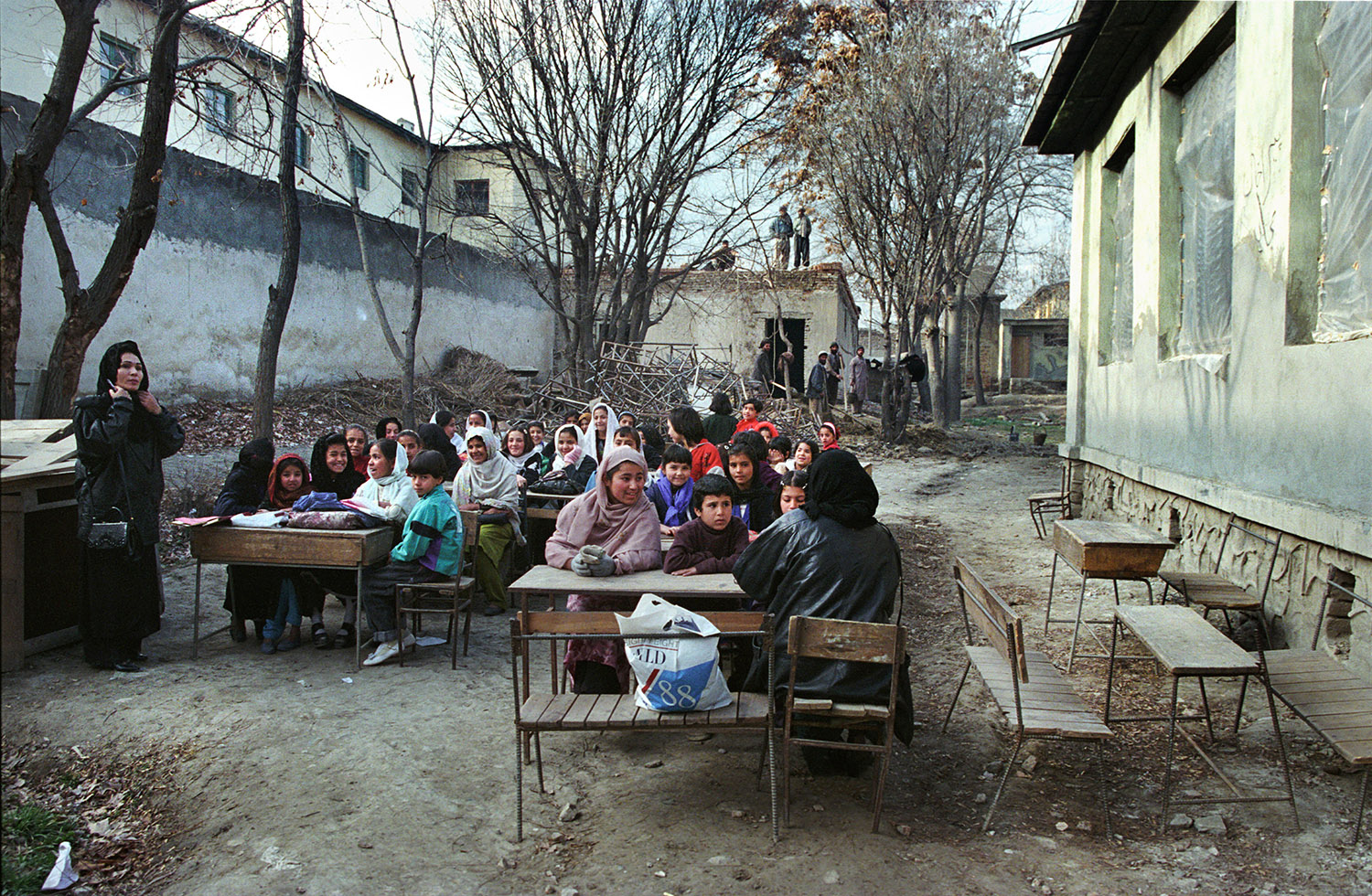 Au Lycée Malalai de Kaboul. Les enfants sont assis, la classe se fait à l'extérieur, dans la cour, les bâtiments ont été détruits. La classe est mixte.