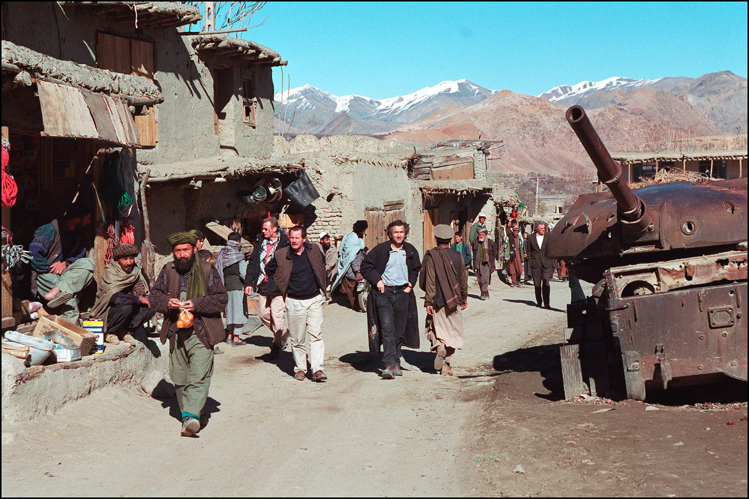 Bernard-Henri Lévy marche dans un village, sur la route de Bamiyan, avec Gilles Hertzog (à droite) et Frédéric Tissot (à gauche), un médecin humanitaire qui a été temporairement chargé d'affaires à l'ambassade de France à Kaboul. Il y a des maisons en terre, un canon abandonné.