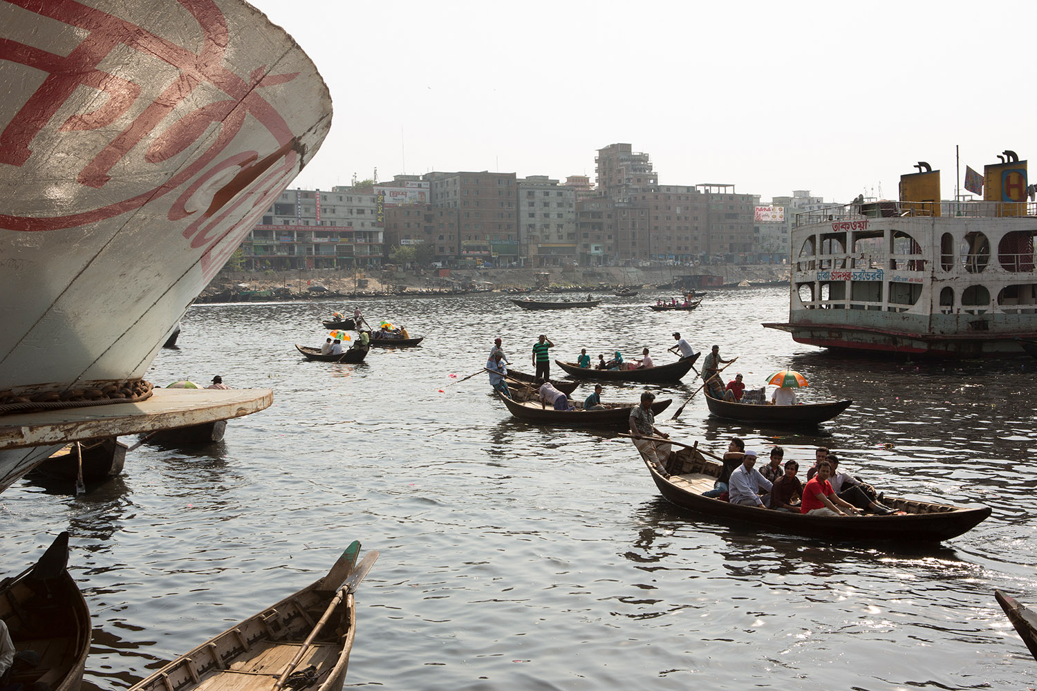 Vue de la rivière Buriganga qui traverse Dacca au Bangladesh. Des petits bateaux de bois sont sur la rivière au milieu de plus gros bateaux.