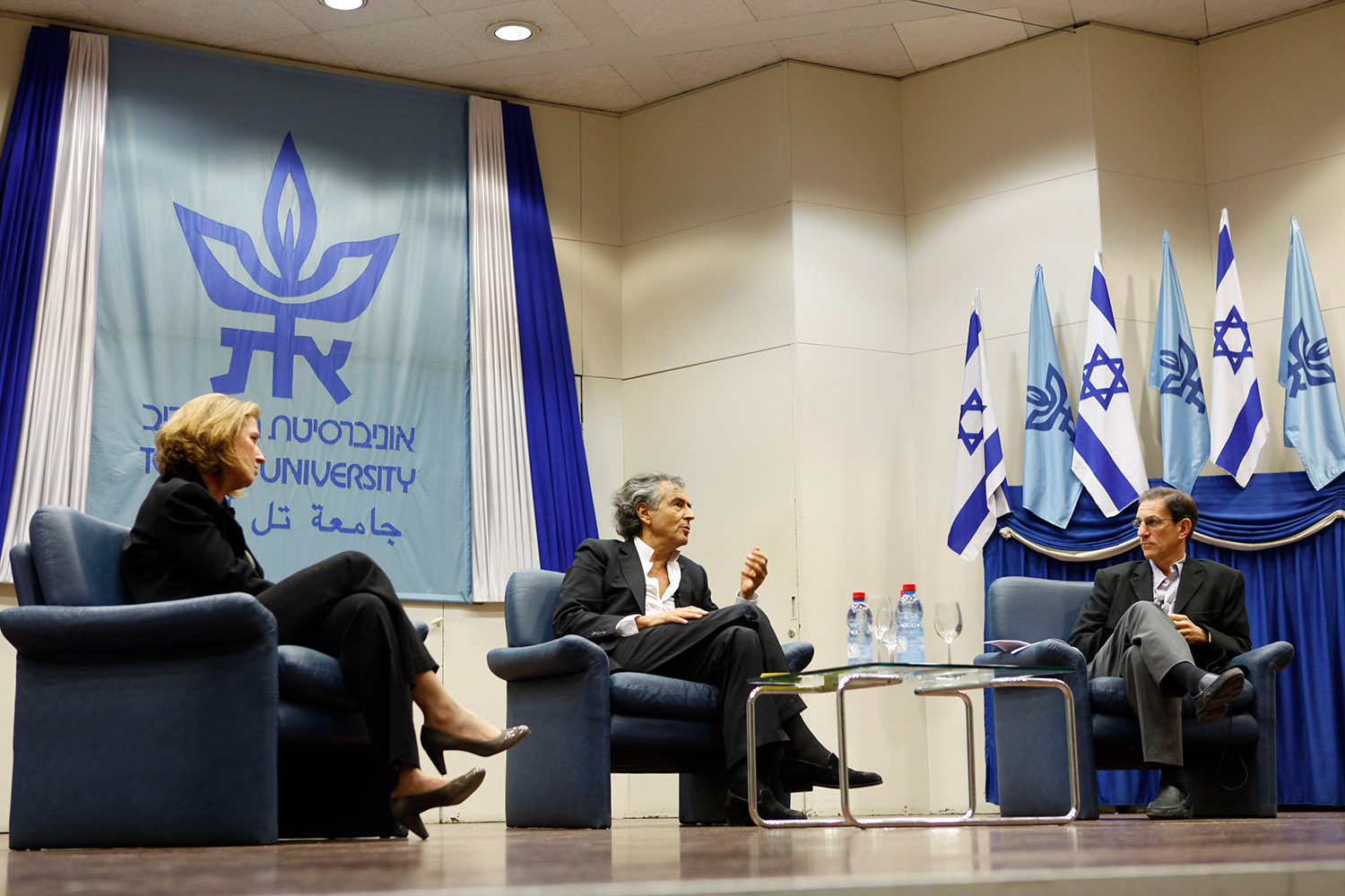 Bernard-Henri Lévy à l'Université de Tel-Aviv. Un homme et une femme sont assis dans des fauteuils à sa droite et à sa gauche. Il y a des drapeaux de l'Université et de l'état d'Israël pendus aux murs.