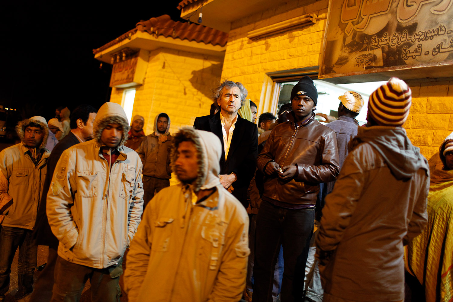Bernard-Henri Lévy dans la ville de Salloum, en Egypte, la nuit, il est entouré par des hommes qui portent des bonnets et des capuches.