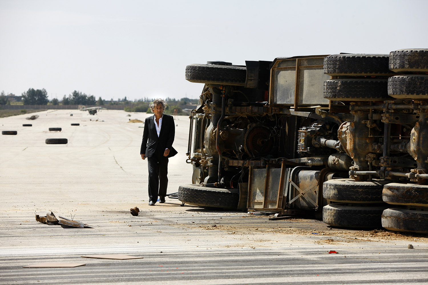 Bernard-Henri Lévy à Labrak, sur la piste de l'aéroport, il marche près d'un camion renversé sur le côté.