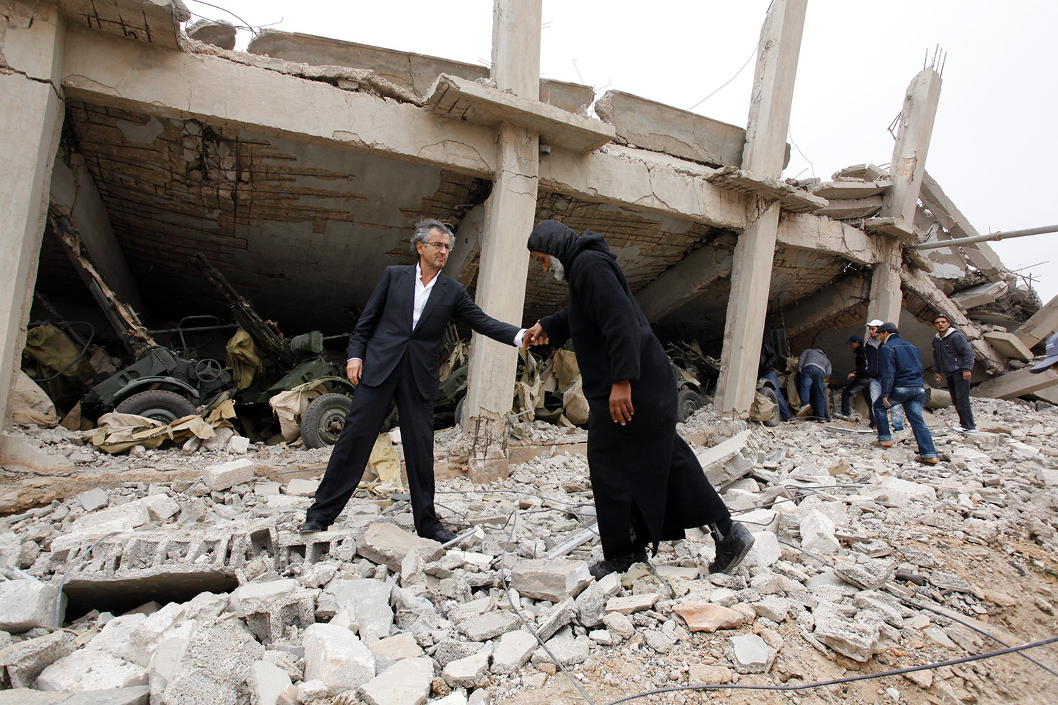 Bernard-Henri Lévy Dans les décombres de la ville de Benghazi en Libye. Il aide un homme à marcher en lui tendant le bras.