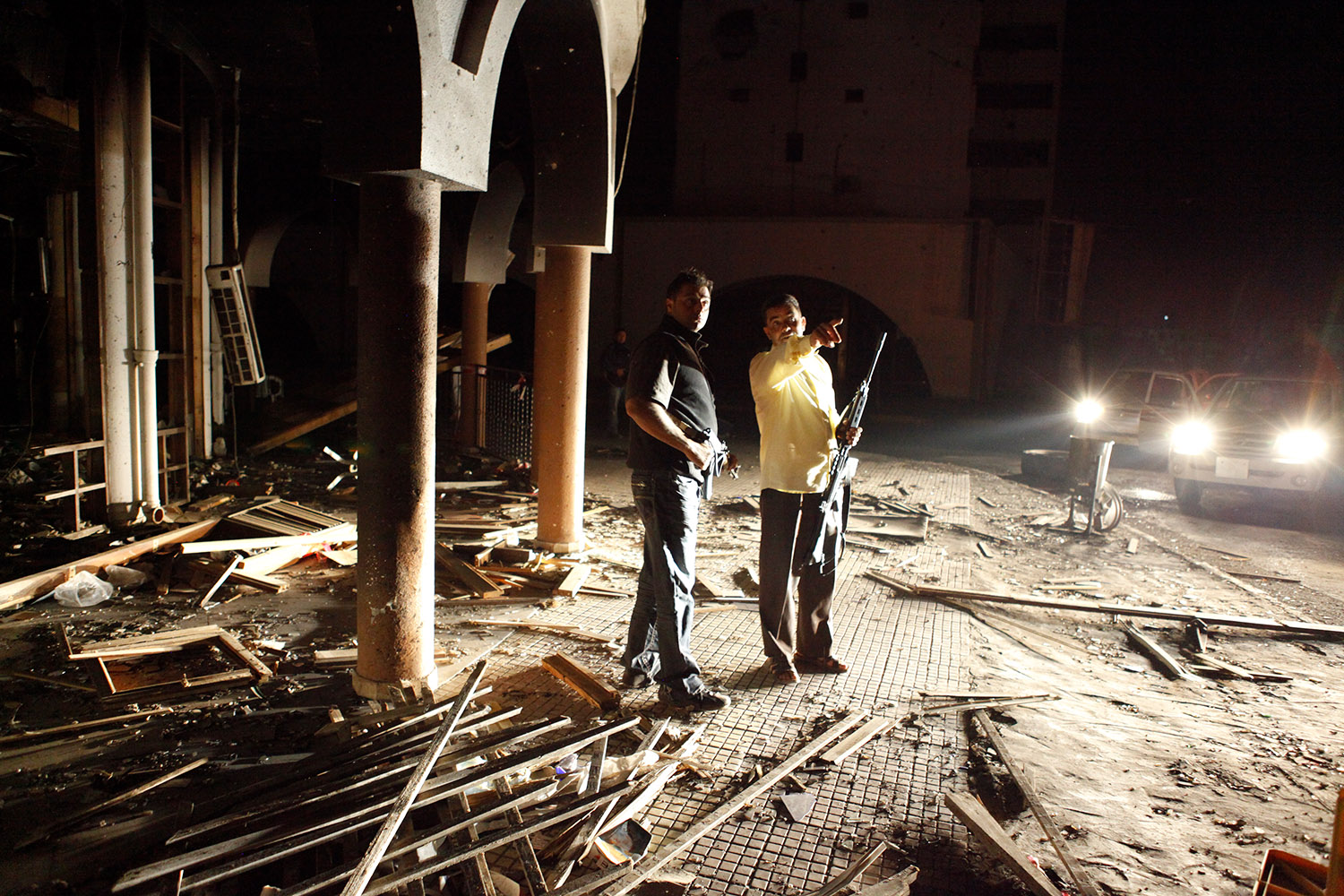 Les phares des voitures éclairent un bâtiment détruit, et ses décombres. au milieu, il y a deux hommes armés.