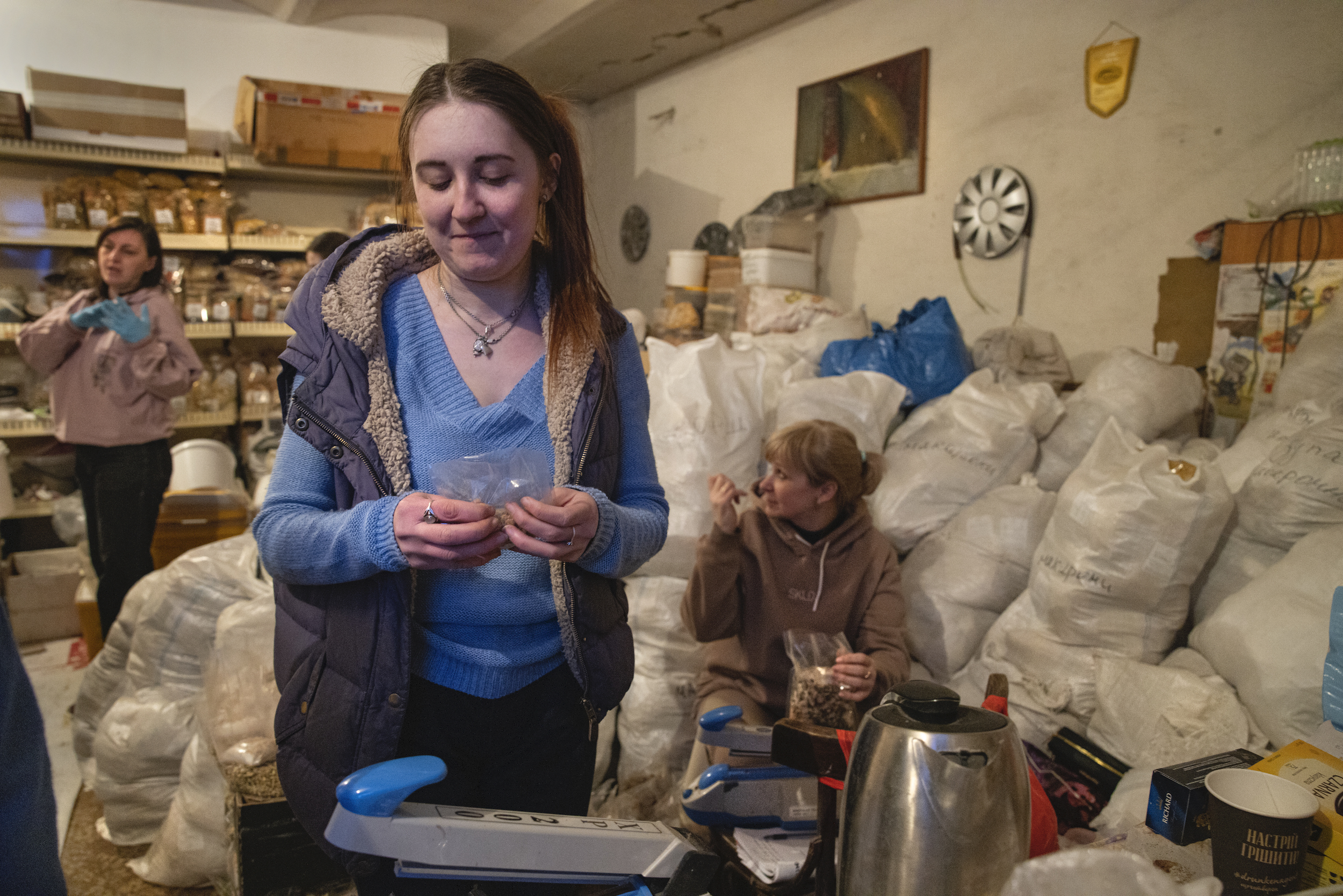 Une pièce avec des femmes qui mettent en ordre des sacs d'alimentations provenant de l'aide humanitaire envoyée par l'étranger à l'Ukraine.La scène se passe dans ce qui semble être un sous-sol.