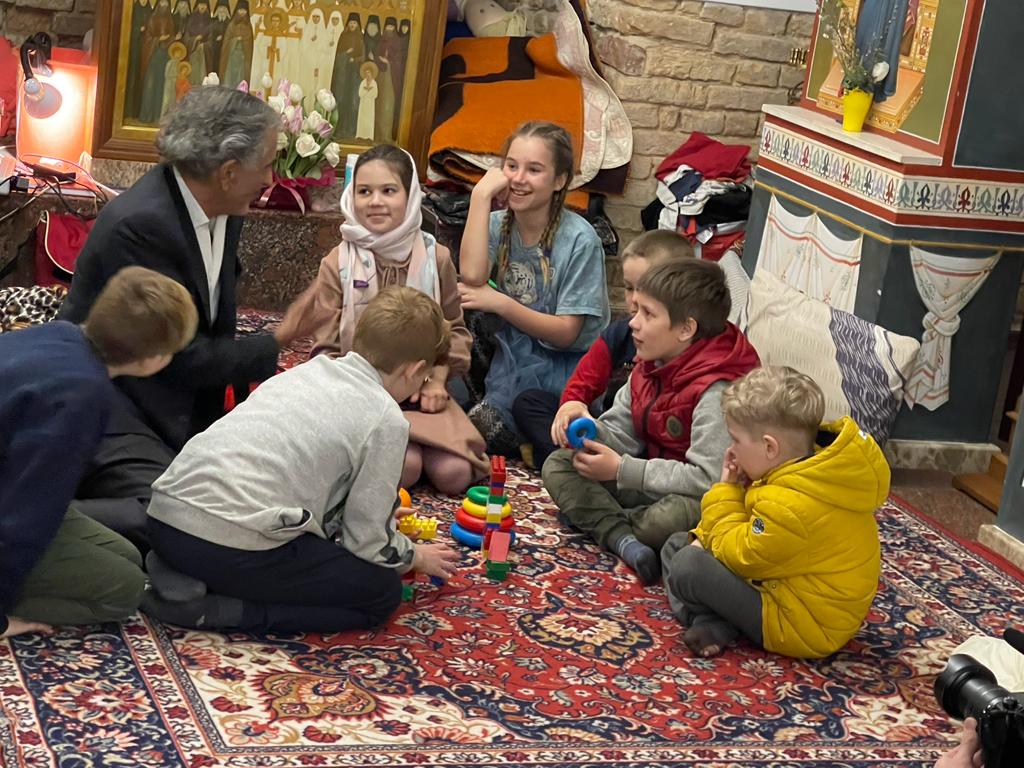 Bernard-Henri Lévy avec des enfants dans un bunker à Kiev. Ils sont assis par terre sur un tapis. On voit des jeux et des icônes religieuses orthodoxes.