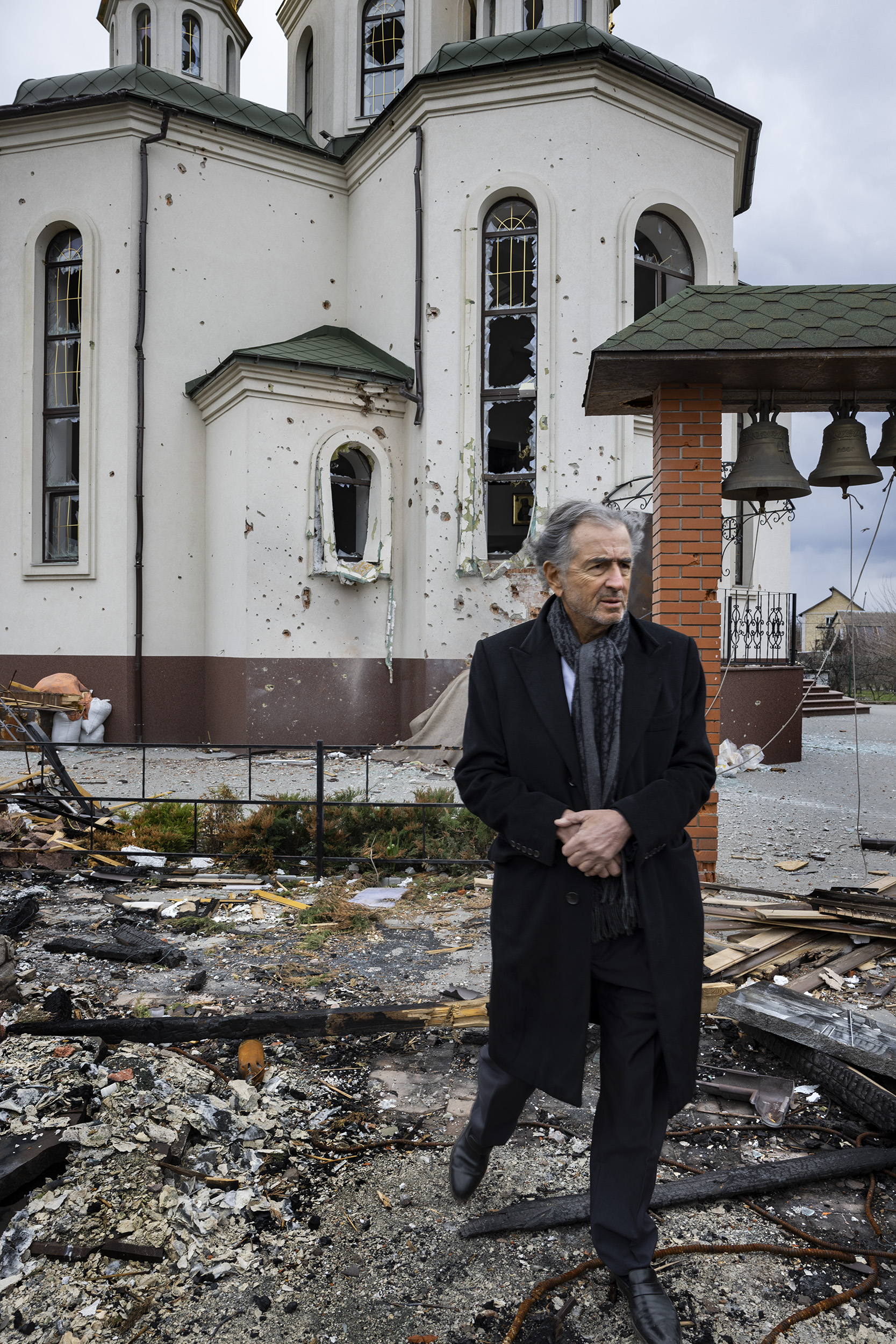 Bernard-Henri Lévy sur les lieux de la guerre en Ukraine, dans un village près de Kiev. Il marche dans les débris. Une église orthodoxe au fond a été détruit. Les vitraux sont cassés, il y a des impacts de balles sur les murs.