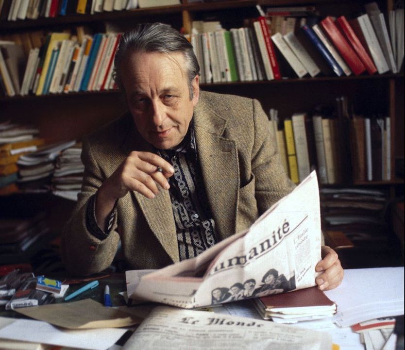 Portrait en couleur de Louis Althusser dans son bureau. Il lit le journal. Derrière lui il y a une bibliothèque.