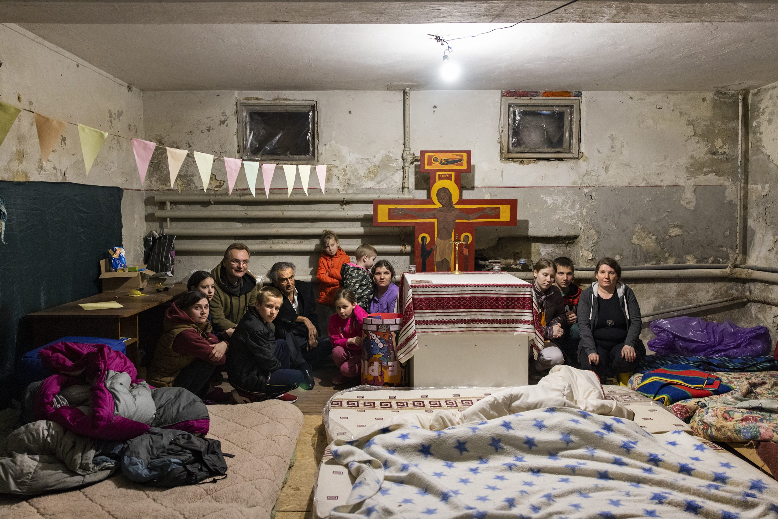 Bernard-Henri Lévy dans les sous-sols un monastère orthodoxe avec une famille. Il y a des matelas au sol, des icônes, une des individus, femmes, enfants et hommes accroupis.