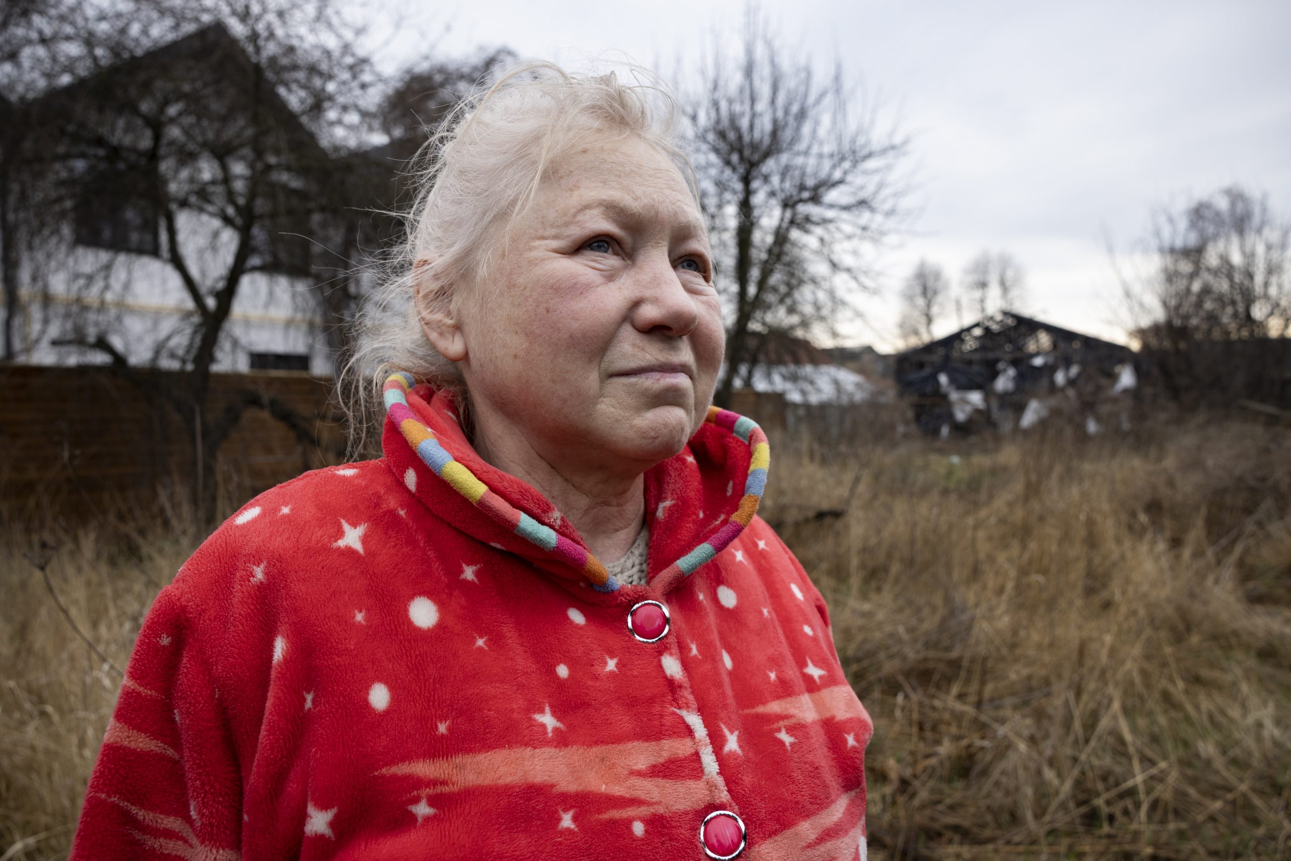 Portrait d'une femme plutôt âgée. Elle a les cheveux blancs et une veste polaire rouge. Derrière elle, les maisons ont été détruites.