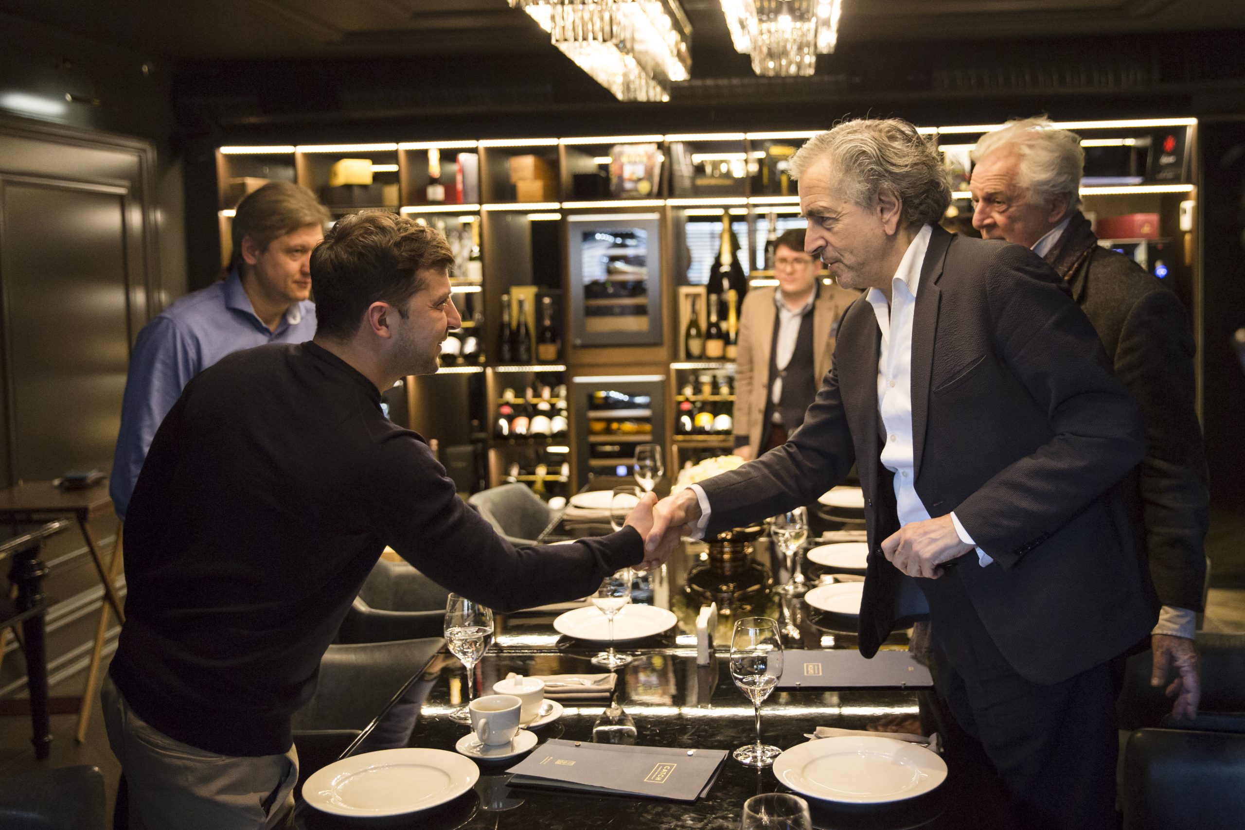 Rencontre entre Bernard-Henri Levy et Volodymyr Zelensky se serrent la main au dessus de la table d'un restaurant.
