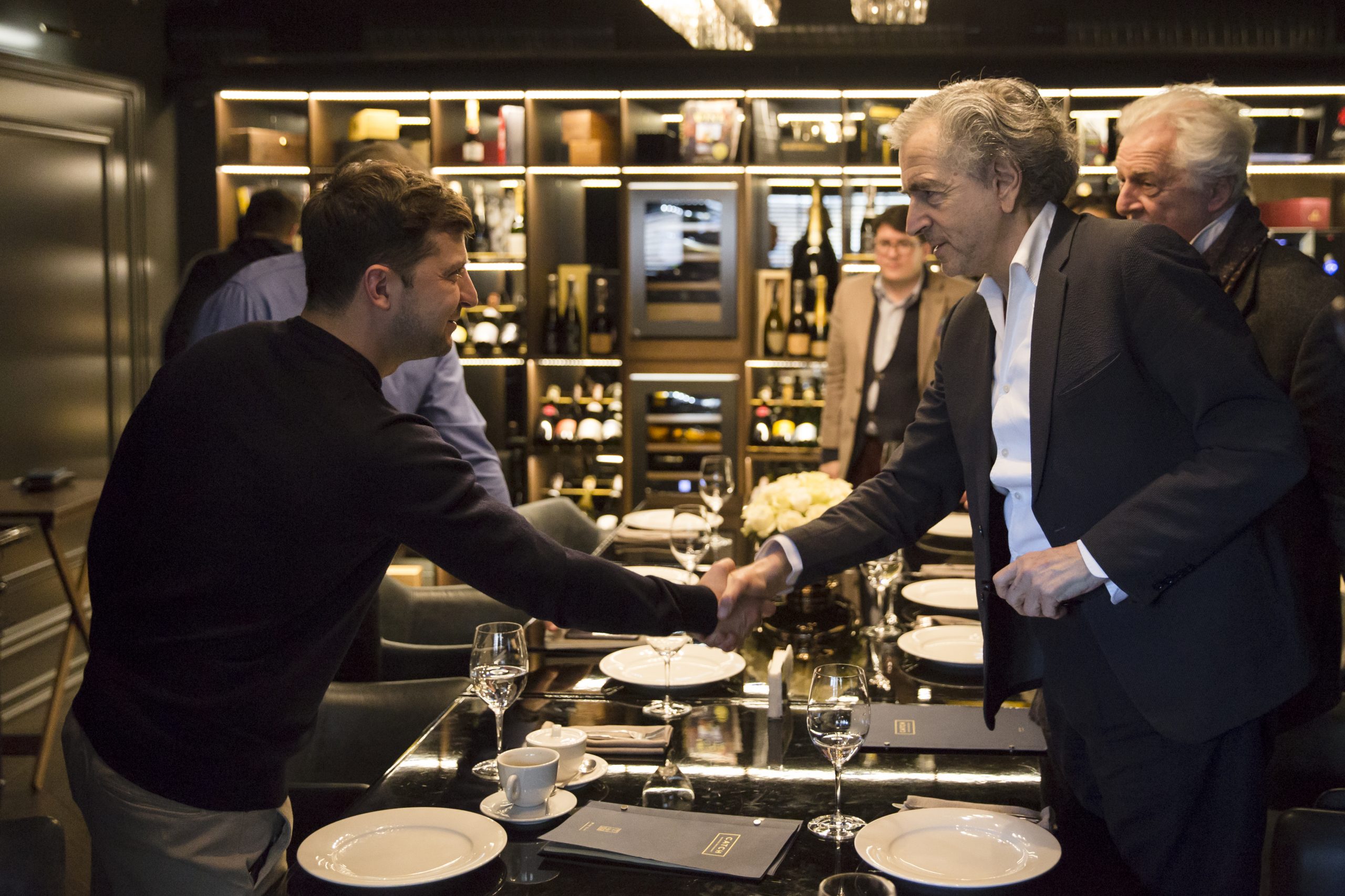 Rencontre entre Bernard-Henri Levy et Volodymyr Zelensky se serrent la main au dessus de la table d'un restaurant.