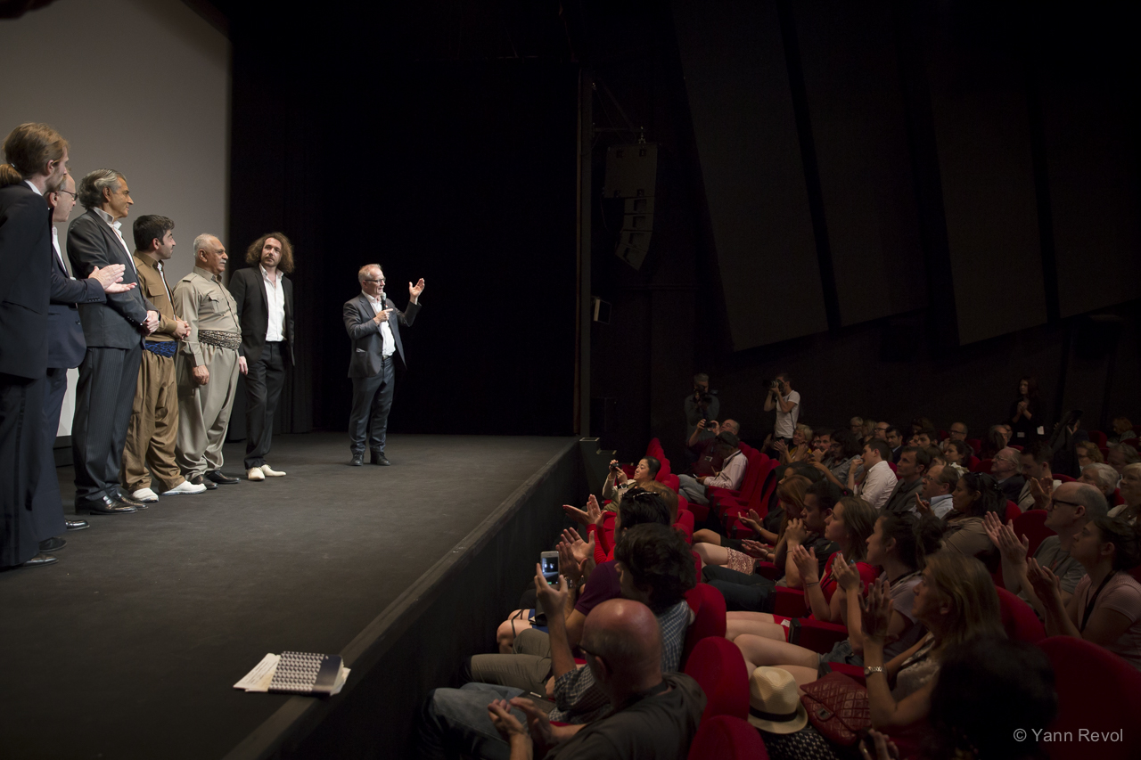 Thierry Frémeaux parle dans un micro sur la scène d'un cinéma. Face à lui la salle est pleine, derrière lui, debout, il y a l'équipe du film de BHL.