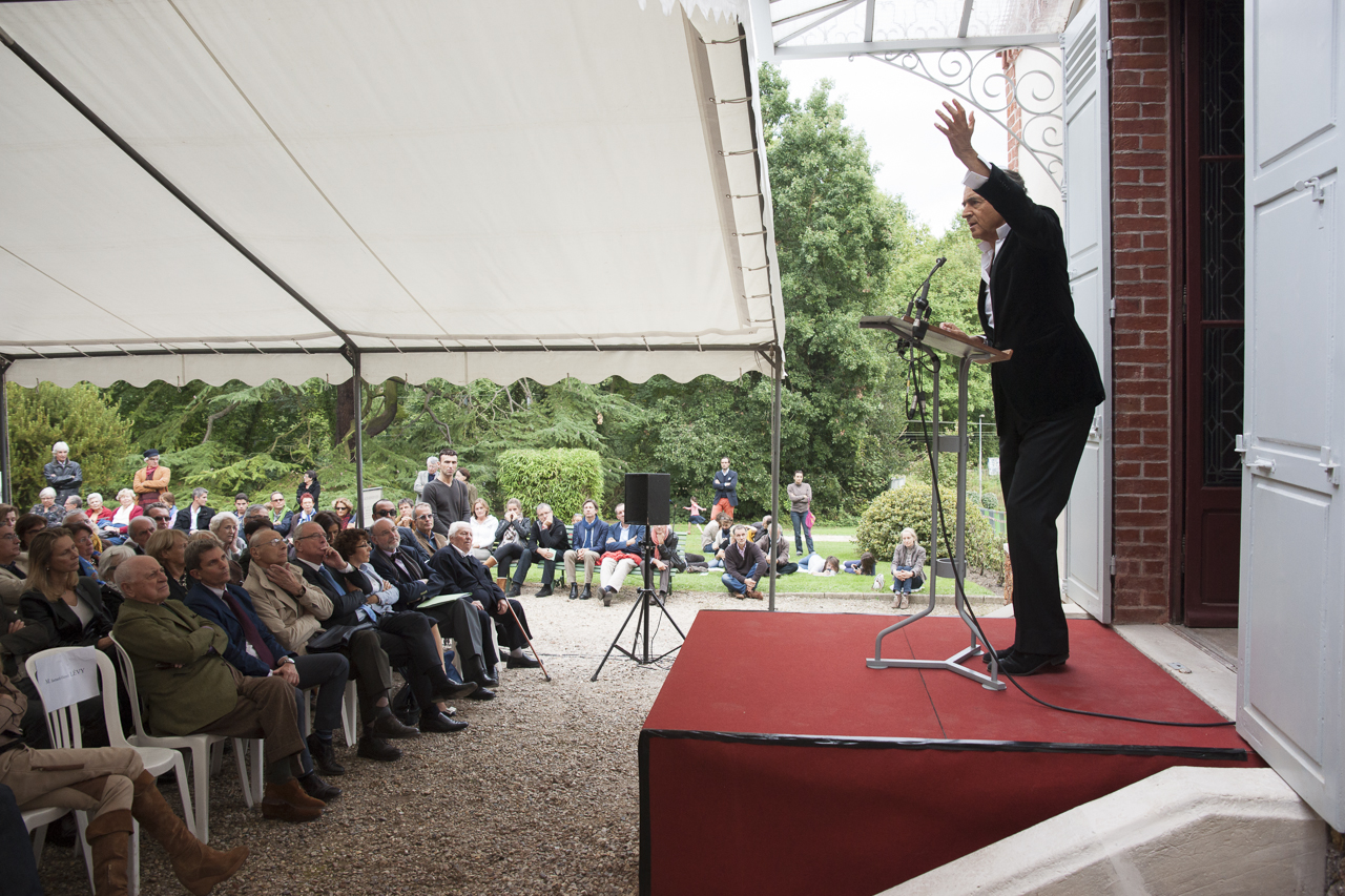 Discours de Bernard-Henri Lévy lors du pèlerinage littéraire à la maison Zola, il est debout sur une scène au seuil de la maison devant un pupitre, face à un public installé sous une tente.