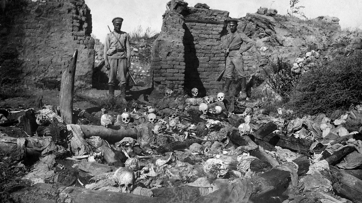 Photo en noir et blanc d'un charnier de victimes arméniennes du génocide dans le village de Sheyxalan. On voit deux soldats devant des restes humains au milieu de ruines.