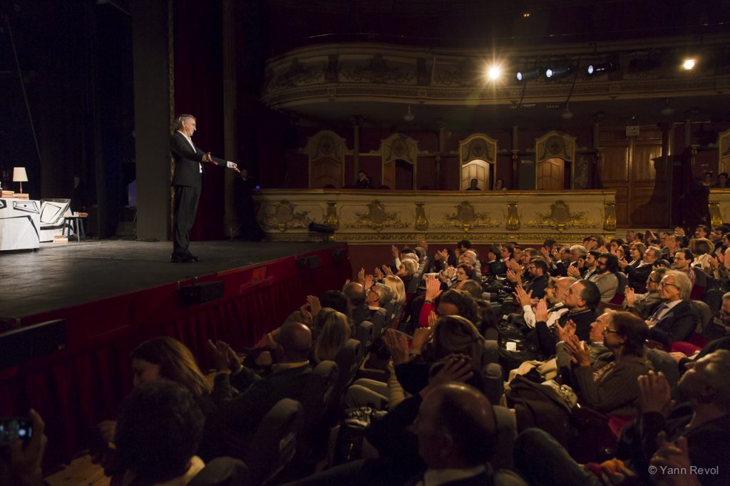 Bernard-Henri Lévy salue le public du Teatro Olympia de Valence, il est seul à l'avant-scène, le public est éclairé.