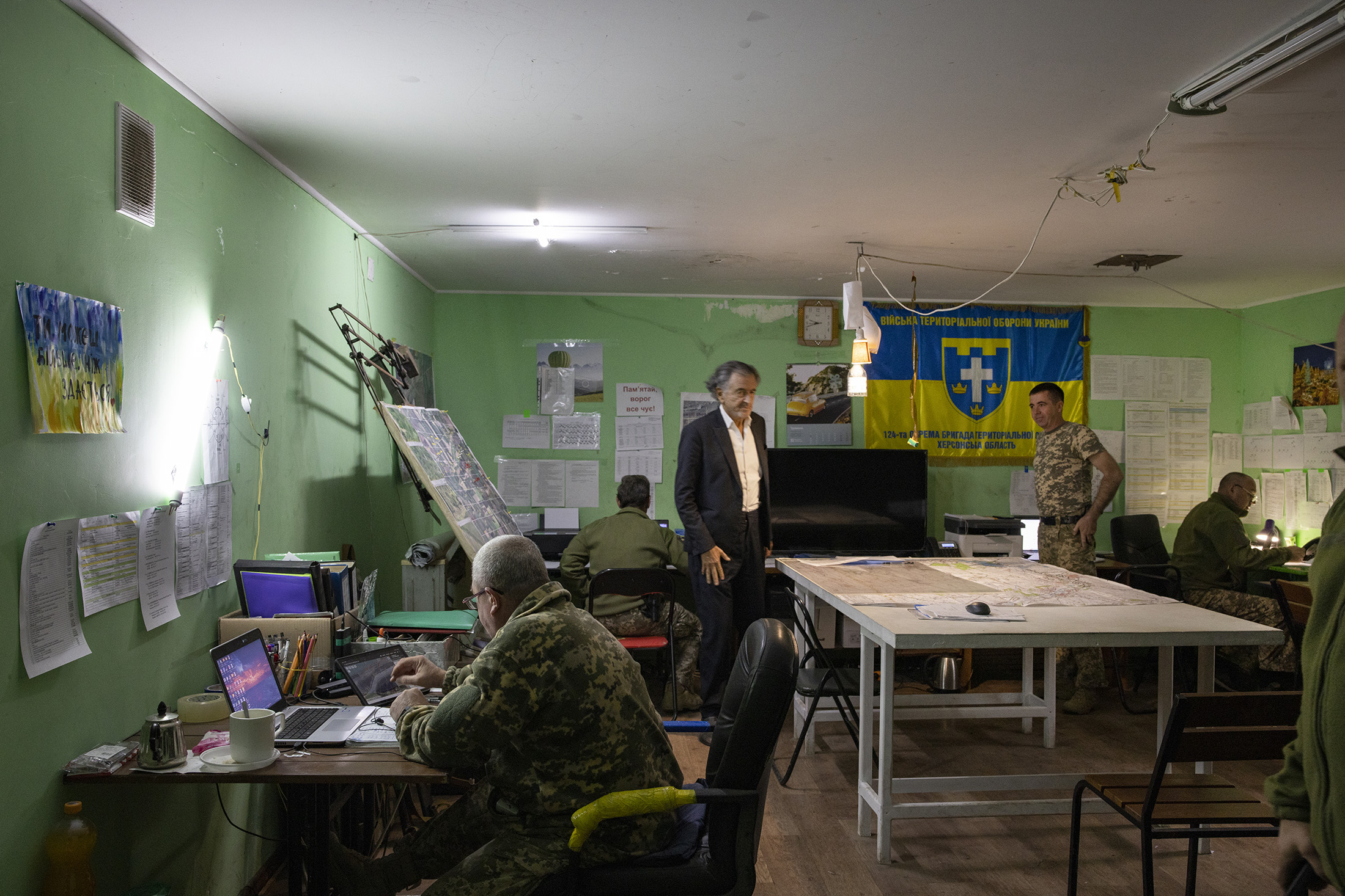 Bernard-Henri Lévy est debout dans une salle où des militaires en uniforme travaillent à des bureaux sur des ordinateurs. Sur les murs sont accrochés des drapeaux aux couleurs de l'Ukraine.