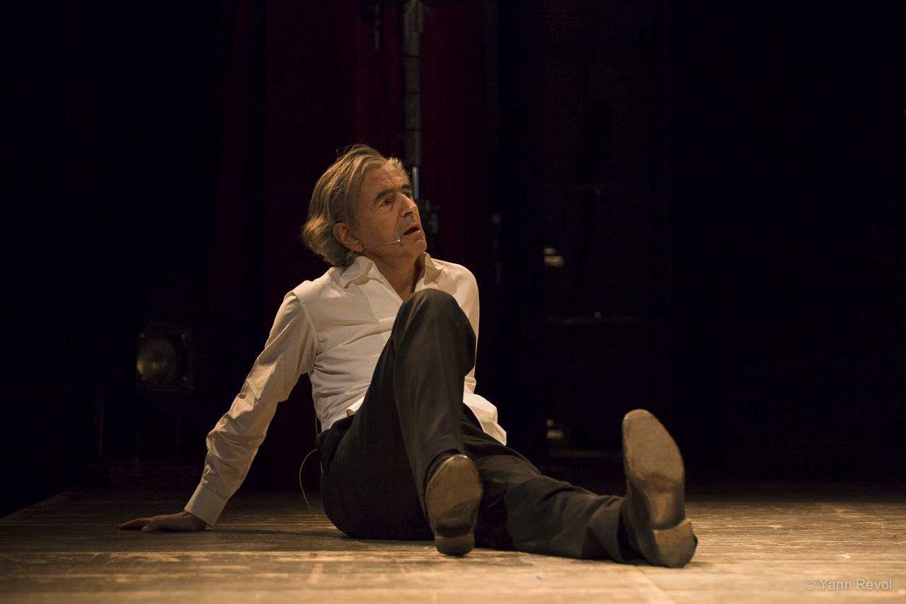 Bernard-Henri Lévy assis sur le sol d'une scène de théâtre.