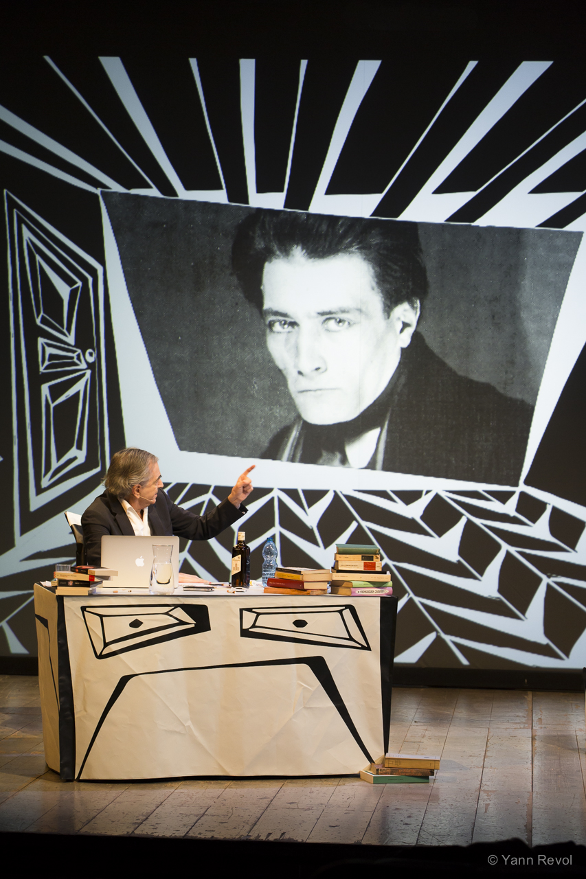 Bernard-Henri Lévy assis derrière un bureau sur une scène de théâtre. Il point un écran au fond du décor qui montre un portrait d'Antonin Artaud.
