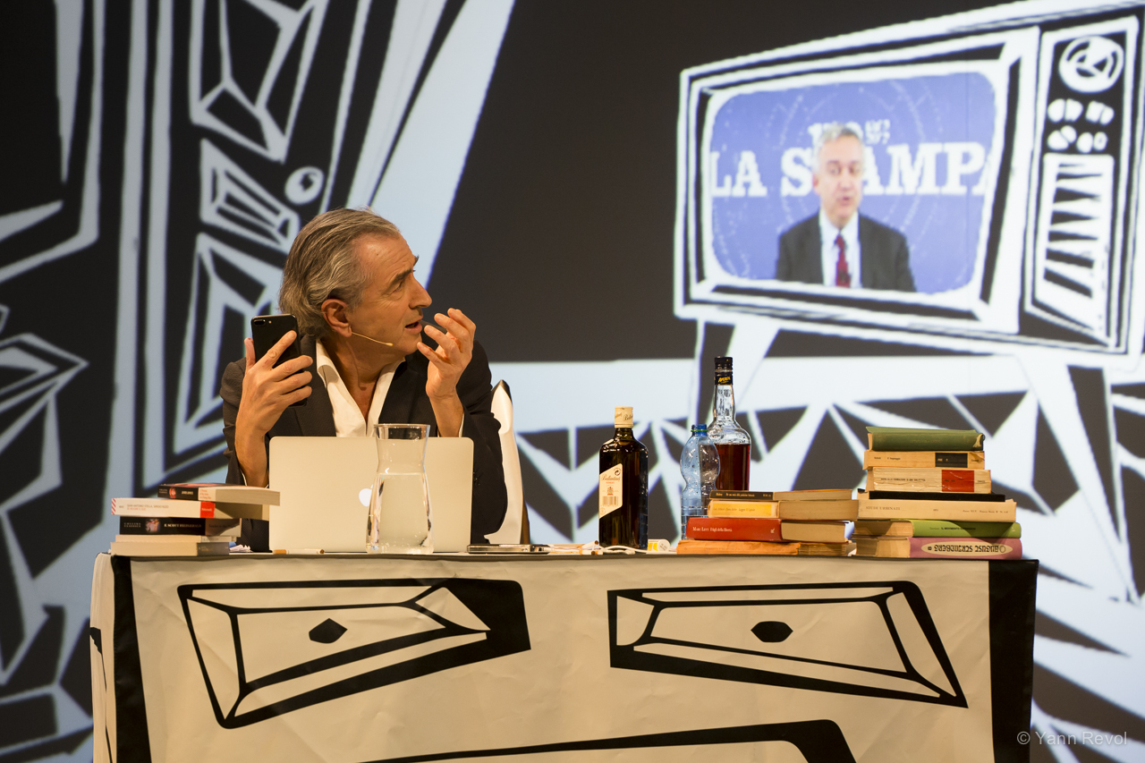 Bernard-Henri Lévy seul en scène assis derrière un bureau sur lequel il y a des livres, un ordinateur et des boissons.