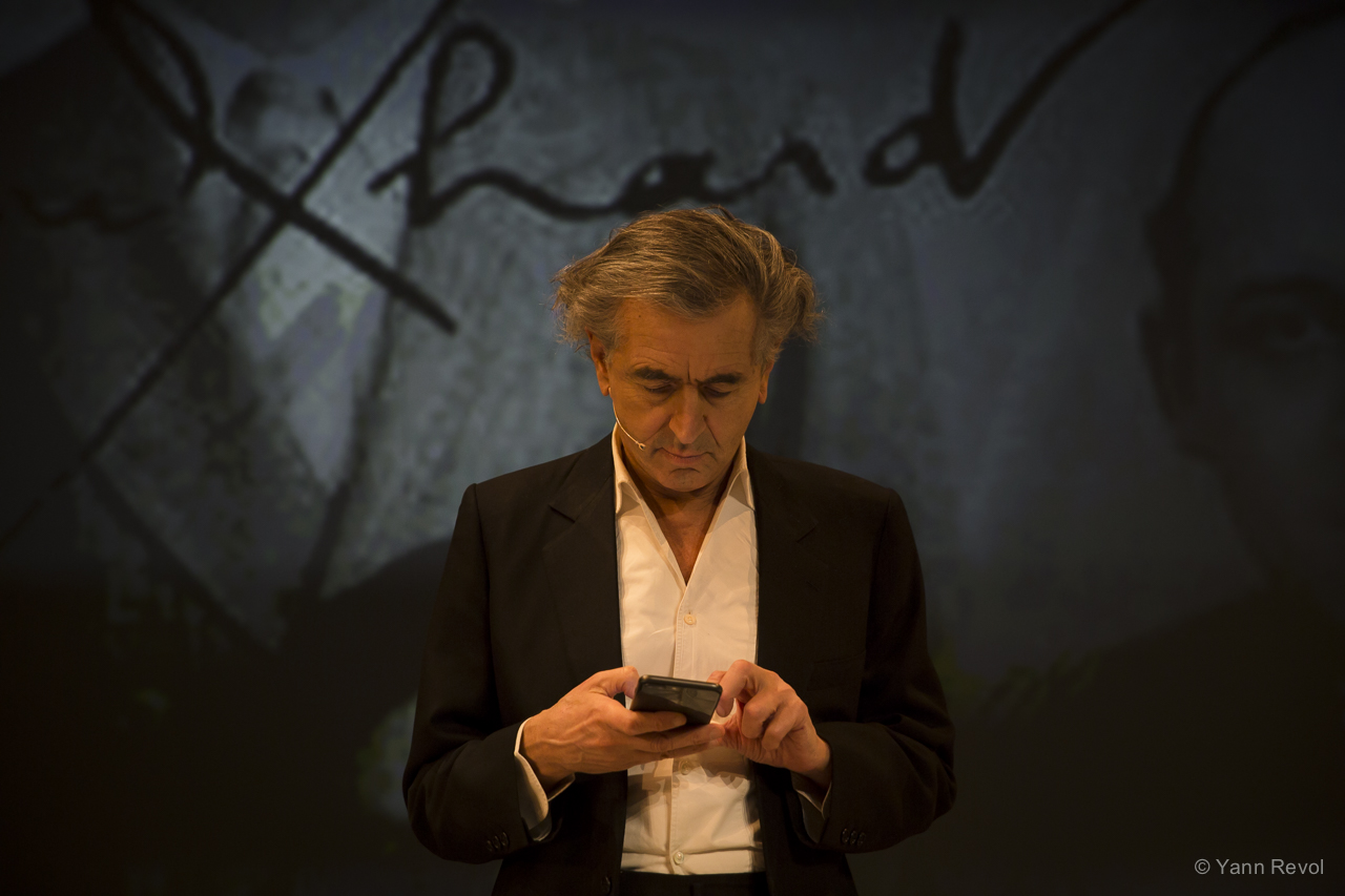 Bernard-Henri Lévy donne une représentation de sa pièce, il est seul en scène, debout, et consulte son téléphone.