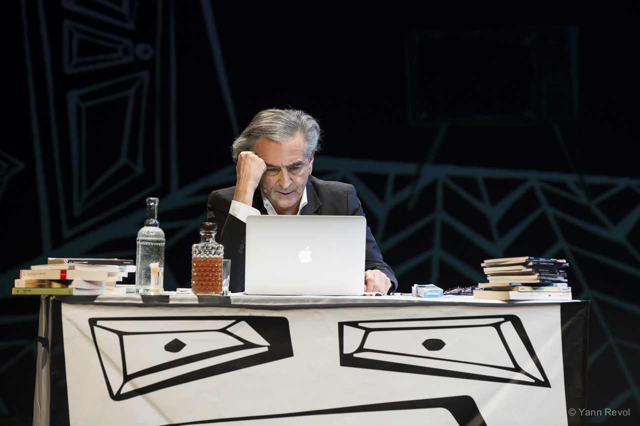 Bernard-Henri Levy interprète son texte de combat « Looking for Europe ». Il est assis, sur scène, derrière un bureau sur lequel il y a un ordinateur portable et des livres.