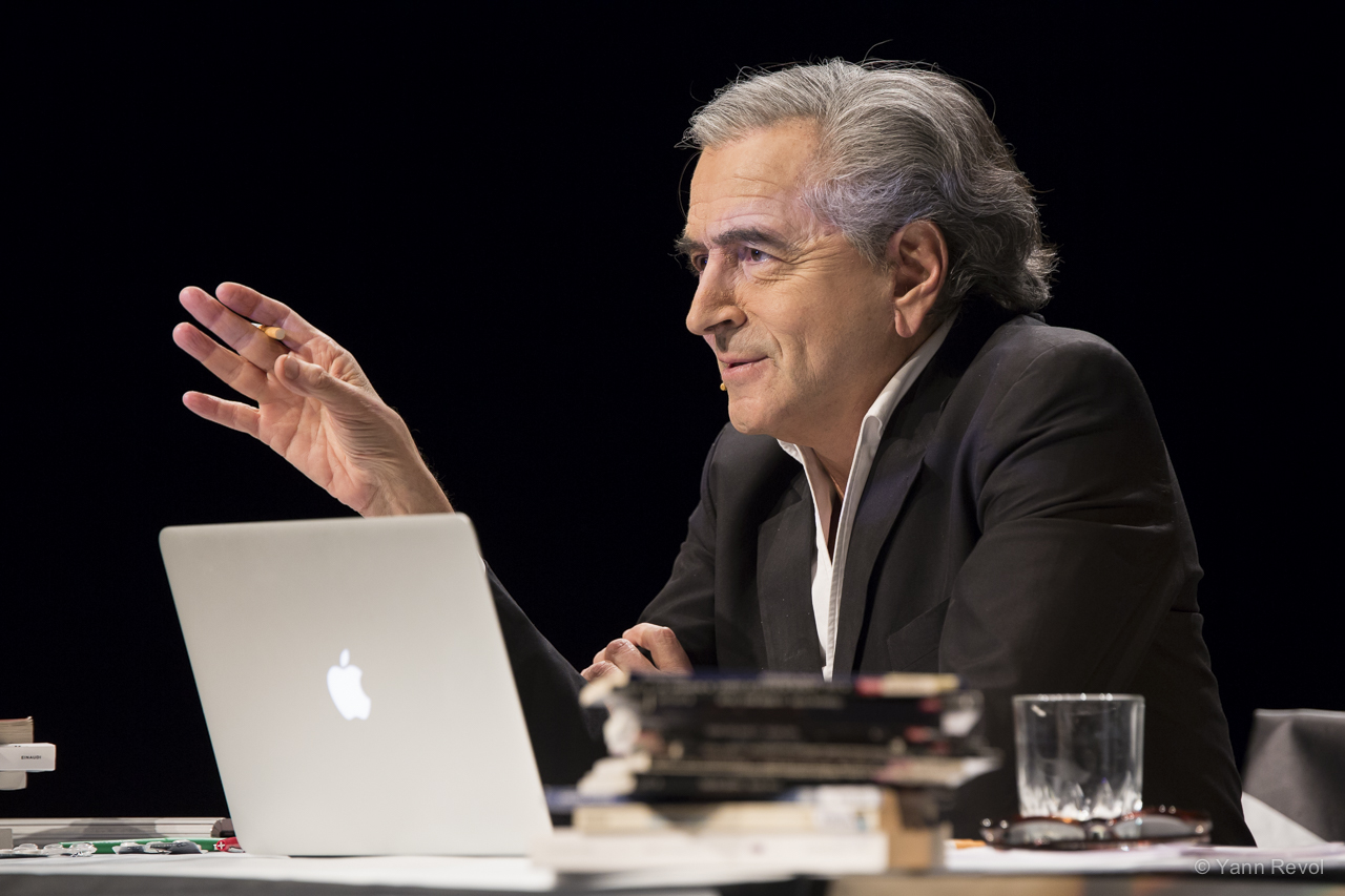 Bernard-Henri Levy interprète son texte de combat « Looking for Europe ». Il est assis, sur scène, derrière un bureau sur lequel il y a un ordinateur portable et des livres.
