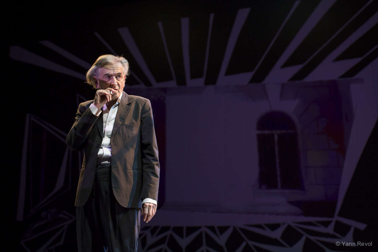 Portrait de Bernard-Henri Lévy sur une scène de théâtre, il tient une cigarette dans la main.