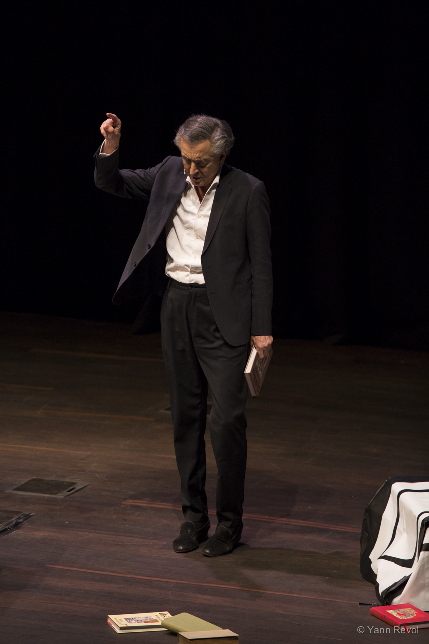 Bernard-Henri Lévy debout sur une scène de théâtre, il tient un livre dans sa main.