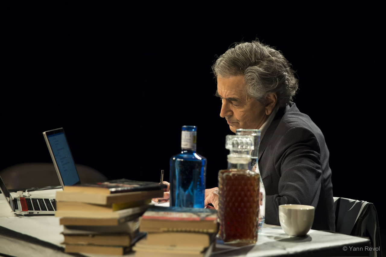 Bernard-Henri Lévy donne une représentation de sa pièce, il est seul assis derrière un bureau sur lequel il y a des livres, des boissons et un ordinateur.