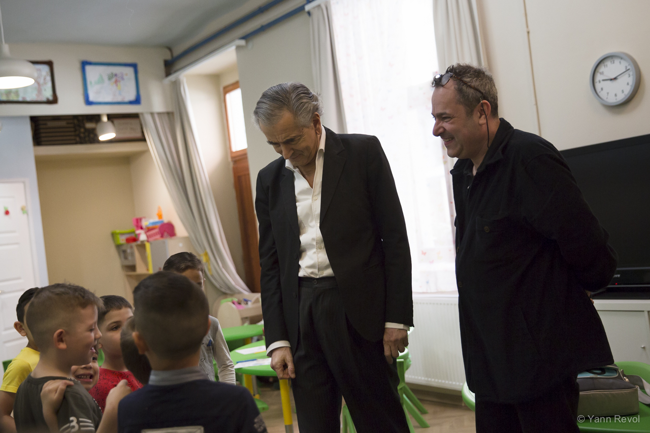 Bernard-Henri Lévy et Balazs Gera visitent l'école pour enfant de l'association caritative Oltalom, ils sont debout dans une salle de classe face à des enfants.