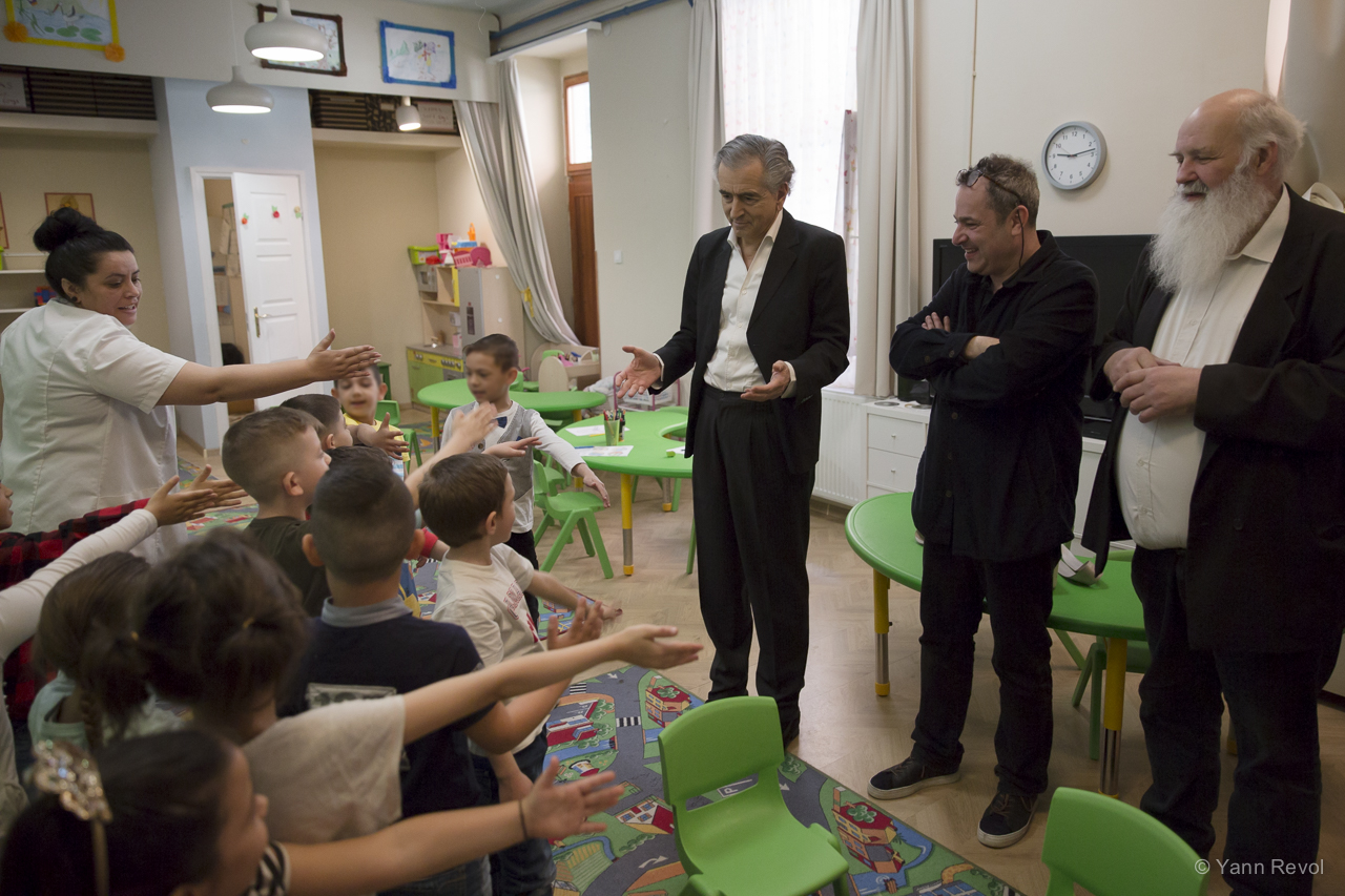 Bernard-Henri Lévy et Balazs Gera visitent avec son créateur Gabor Ivanyi l'école pour enfant de l'association caritative Oltalom, ils sont debout dans une salle de classe face à des enfants.