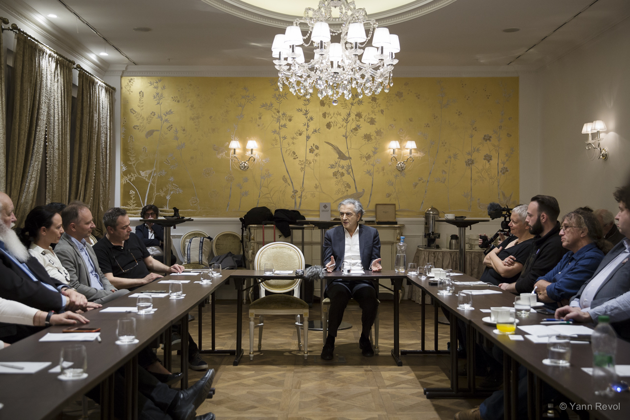 Bernard-Henri Lévy est assis autour d'une table et s'adresse à une douzaine de personnes également assis autour de la table.