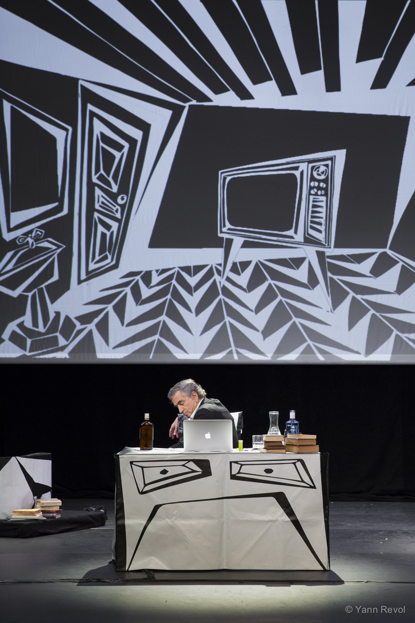 Bernard-Henri Lévy joue sa pièce « Looking for Europe » au Théâtre Antoine à Paris. Il est seul sur scène assis derrière un bureau sur lequel il y a un ordinateur et des livres.