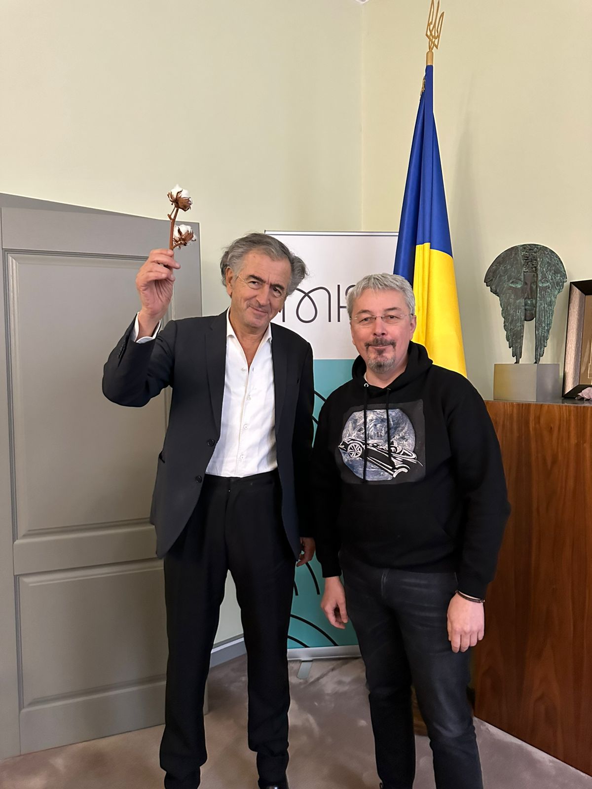 Oleksandr Tkachenko avec Bernard-Henri Lévy. Ils posent devant un drapeau ukrainien. BHL tient une fleur de coton.